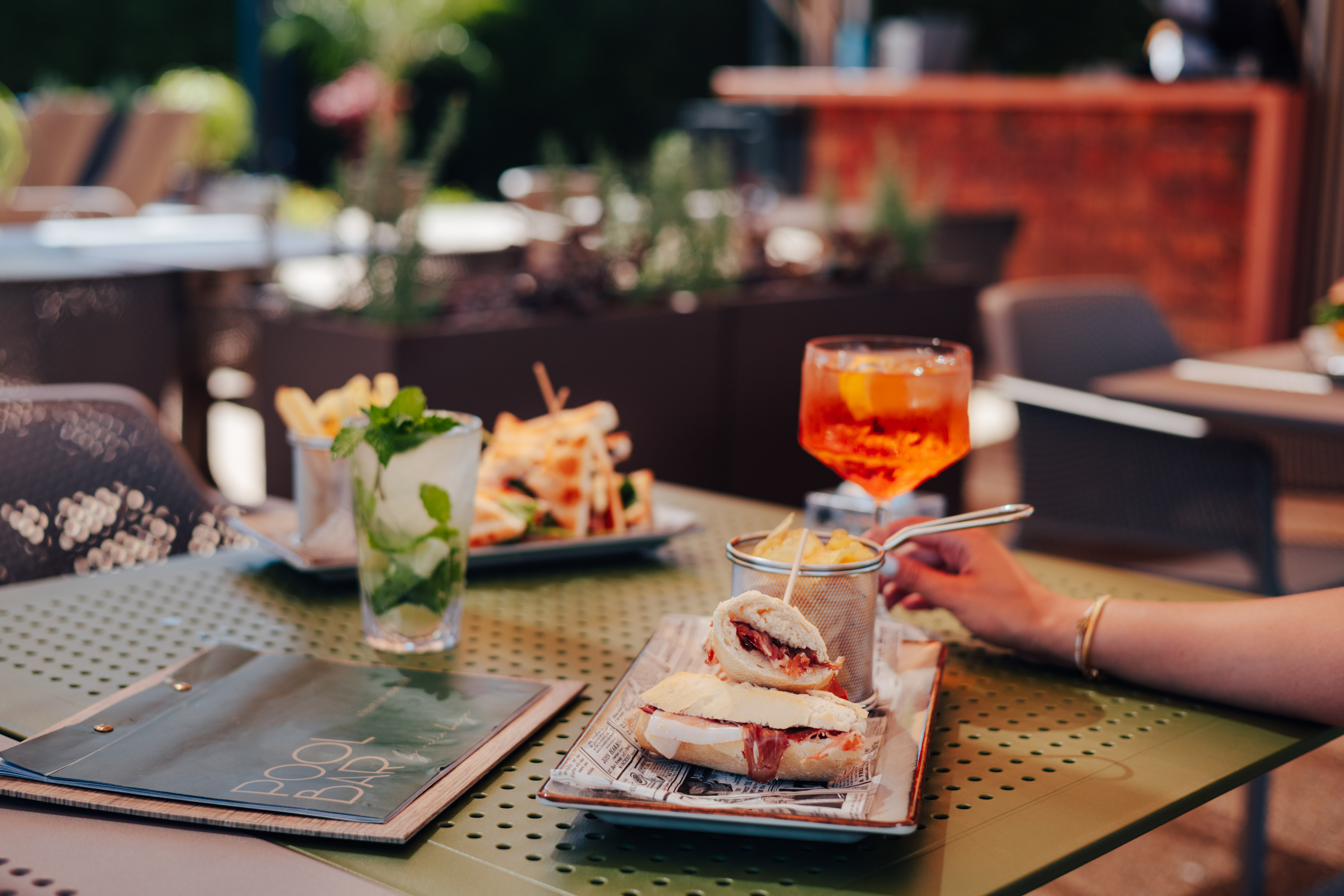 Outdoor patio dining with sandwiches, fries, mojito, and Aperol Spritz.