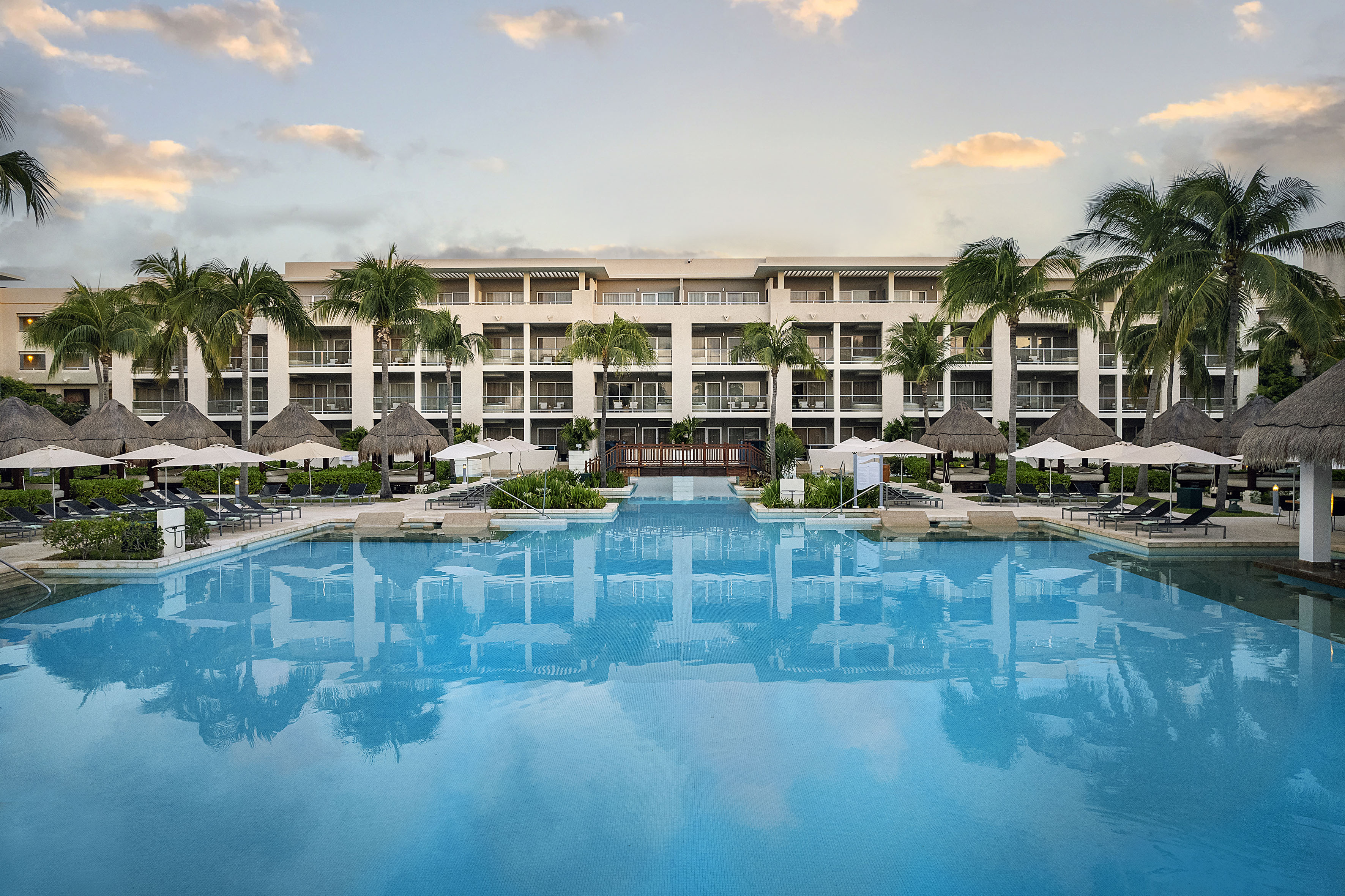 Tropical resort pool with hotel building, palm trees, and thatched umbrellas.