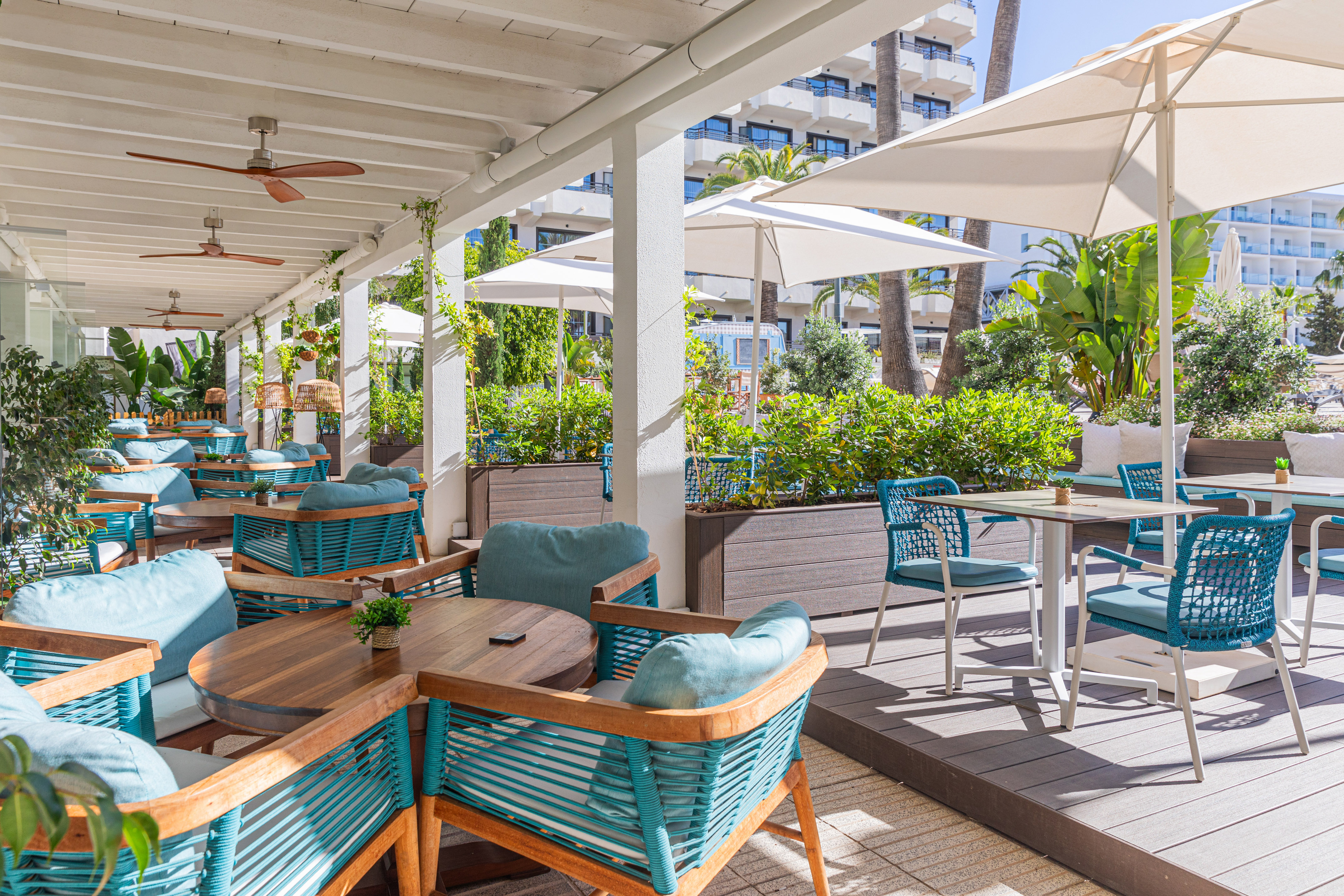 Sunny hotel restaurant terrace with teal seating, wooden tables, and umbrellas.