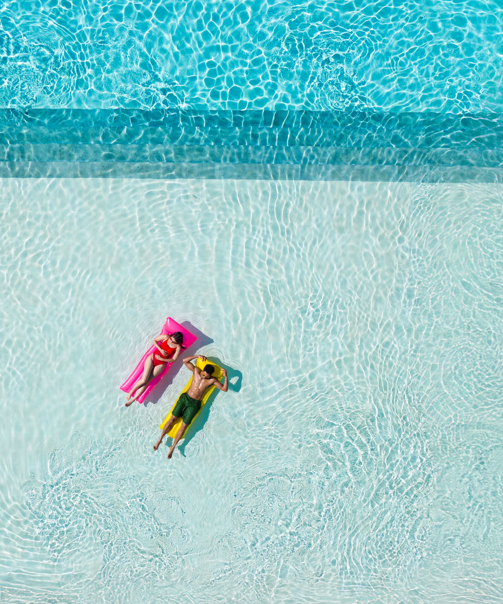 Couple floating on pink and yellow inflatables in a bright blue pool.