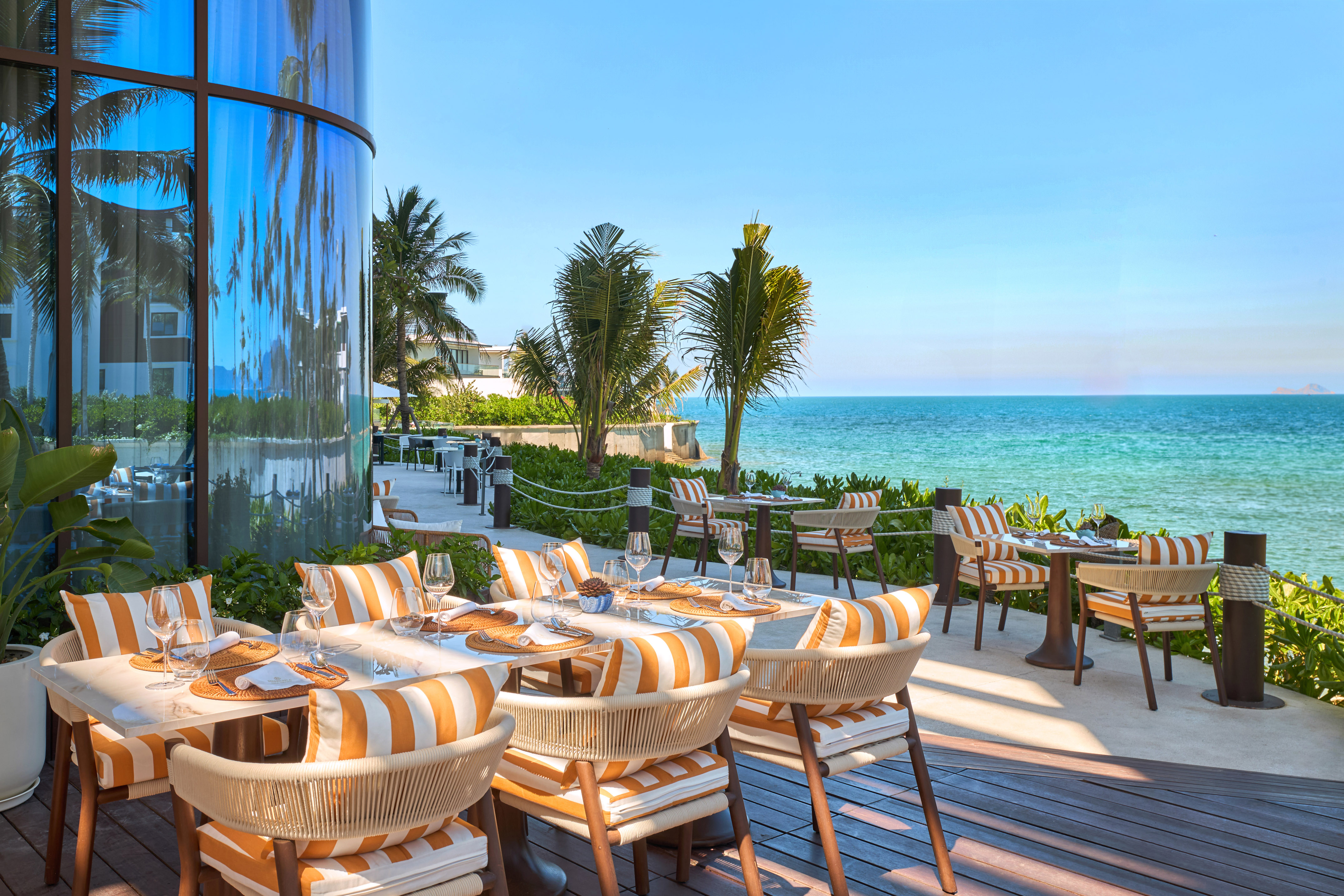 Oceanfront dining patio with striped chairs, palm trees, and blue ocean view.