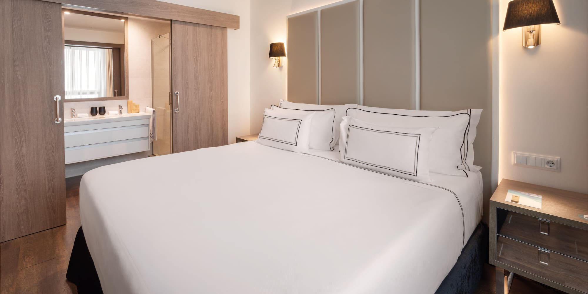 Modern hotel bedroom with white bed, beige headboard, and open bathroom.