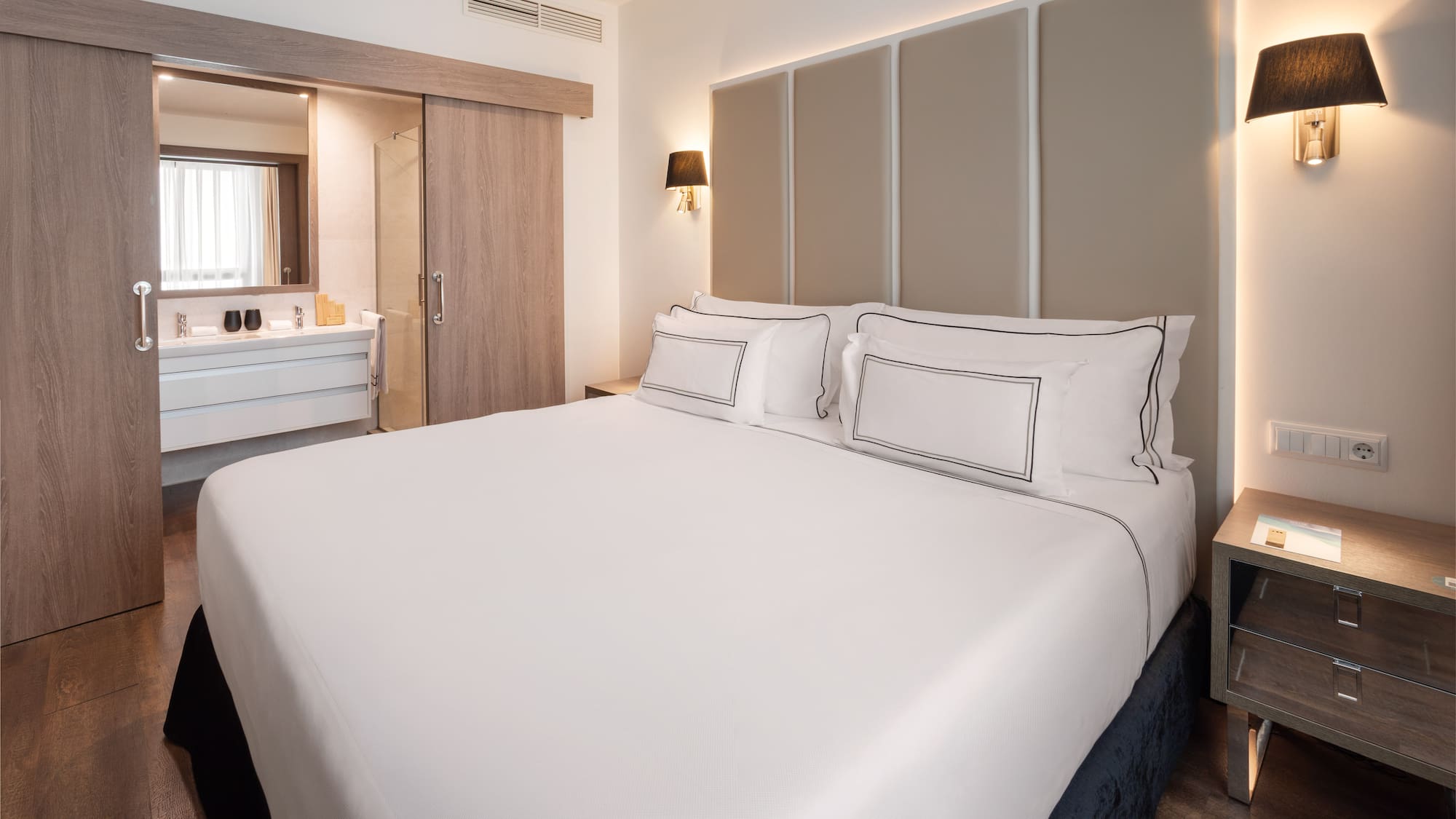 Modern hotel bedroom with white bed, beige headboard, and open bathroom.