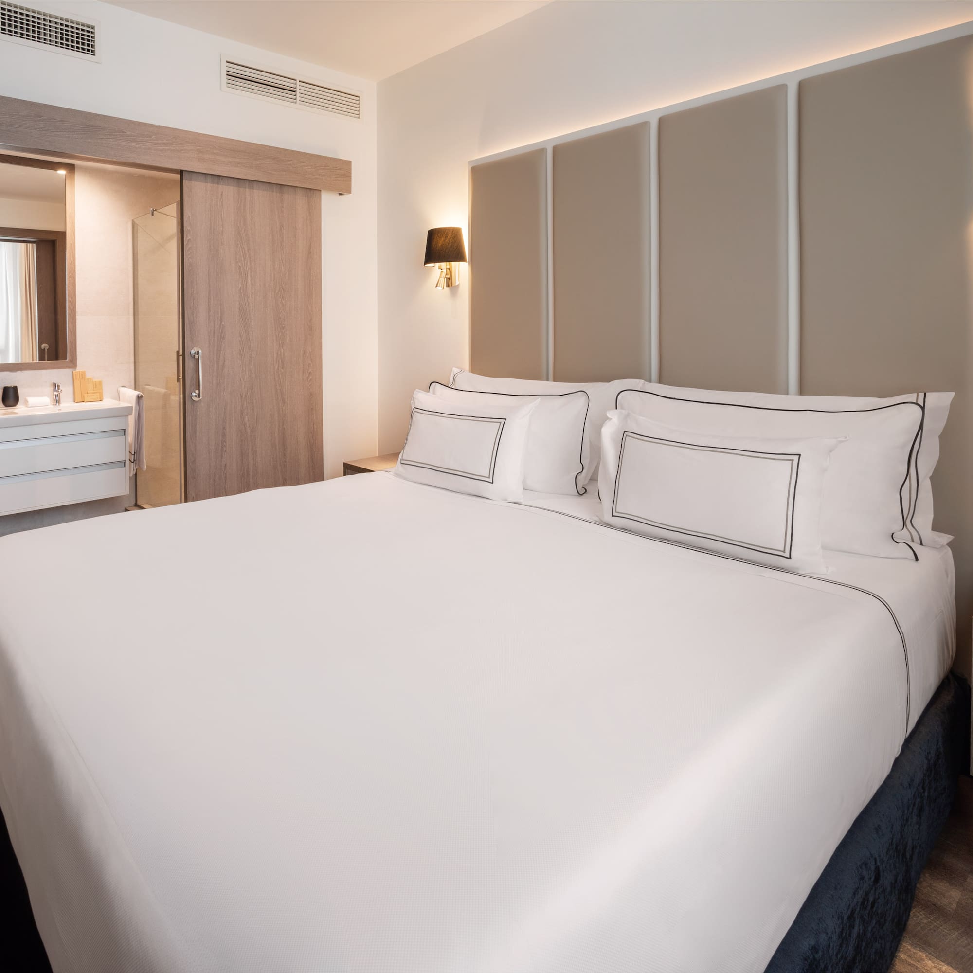Modern hotel bedroom with white bed, beige headboard, and open bathroom.