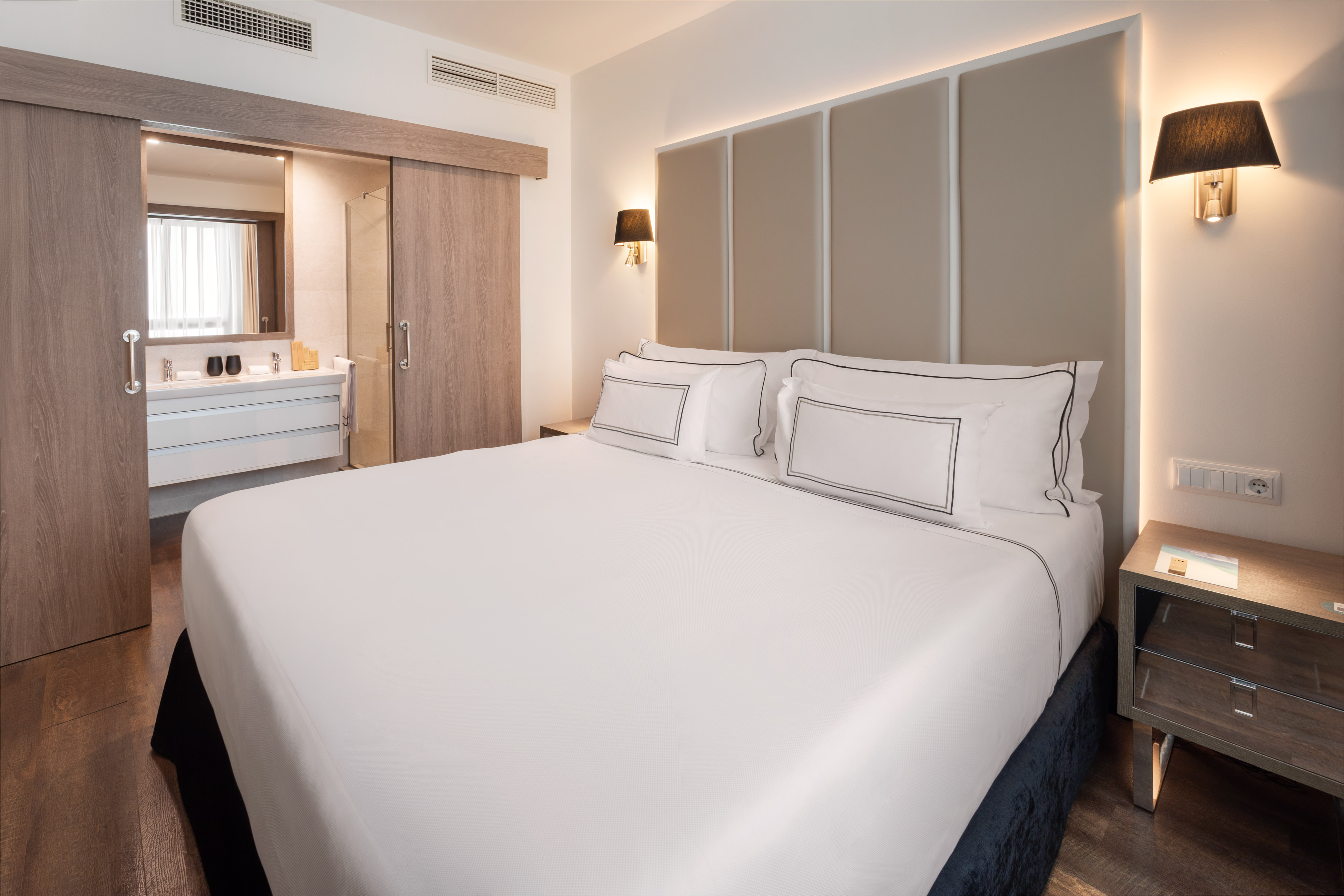 Modern hotel bedroom with white bed, beige headboard, and open bathroom.