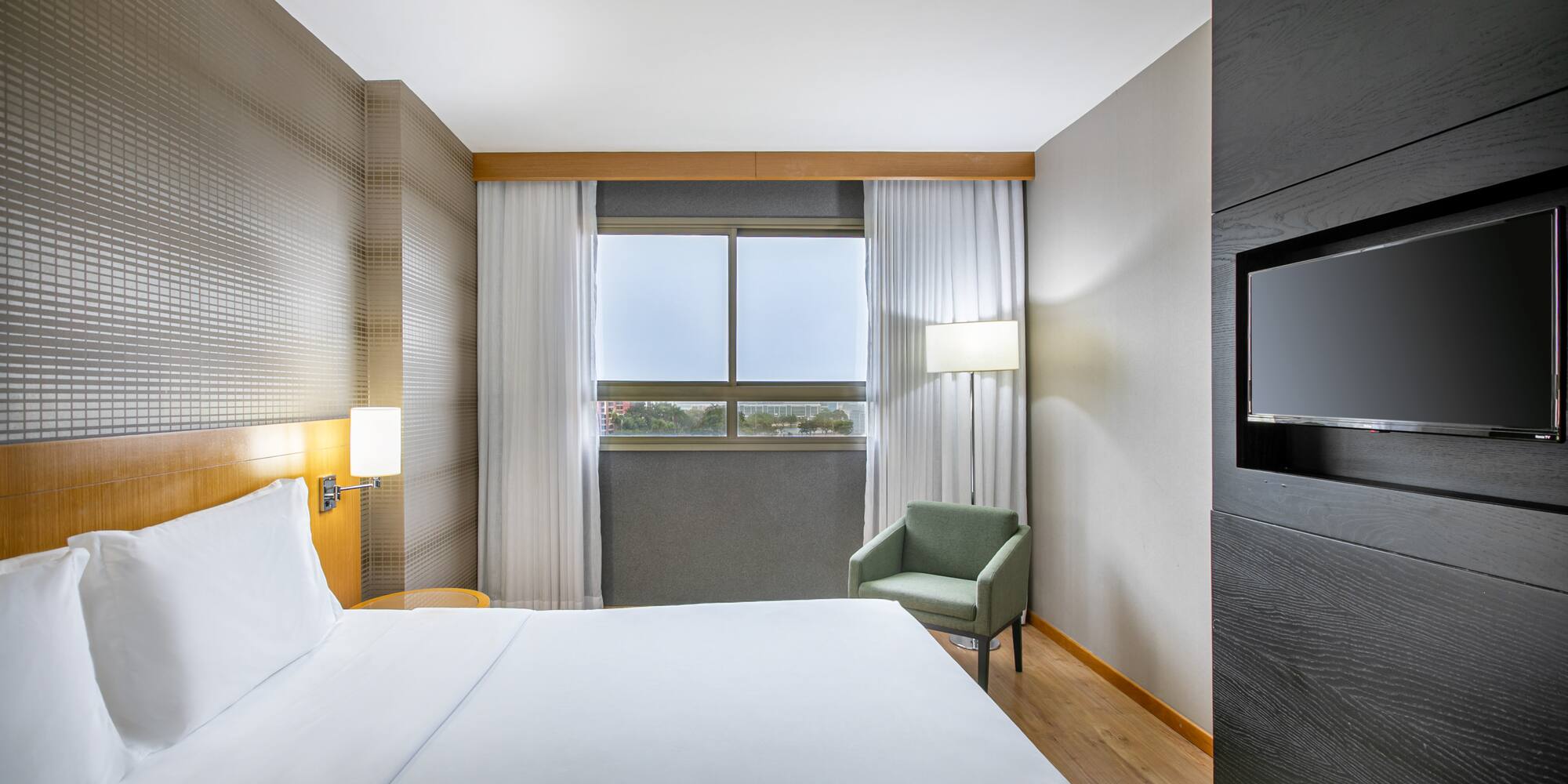 Sleek hotel room with white bed, window, armchair, and mounted flat-screen TV.