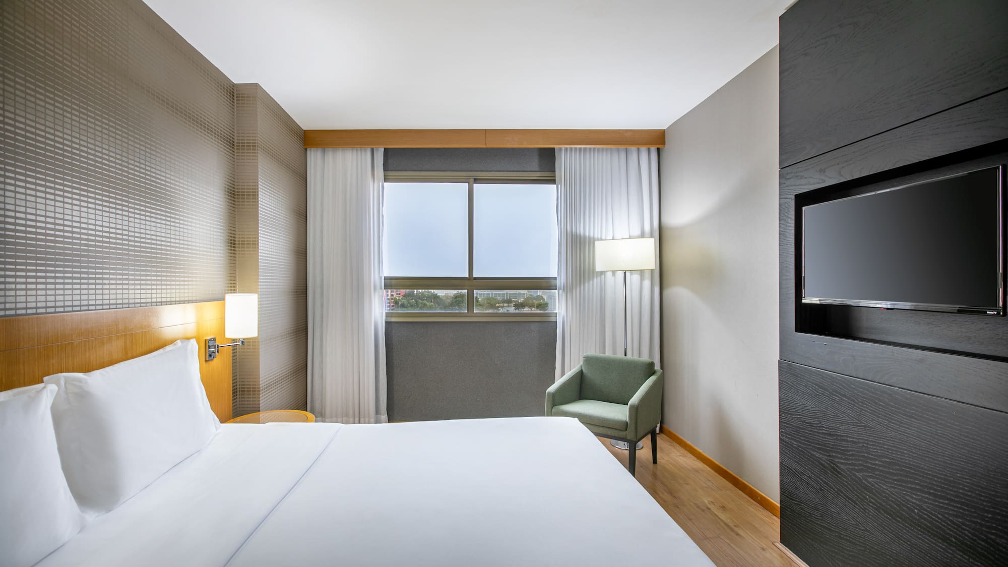 Sleek hotel room with white bed, window, armchair, and mounted flat-screen TV.