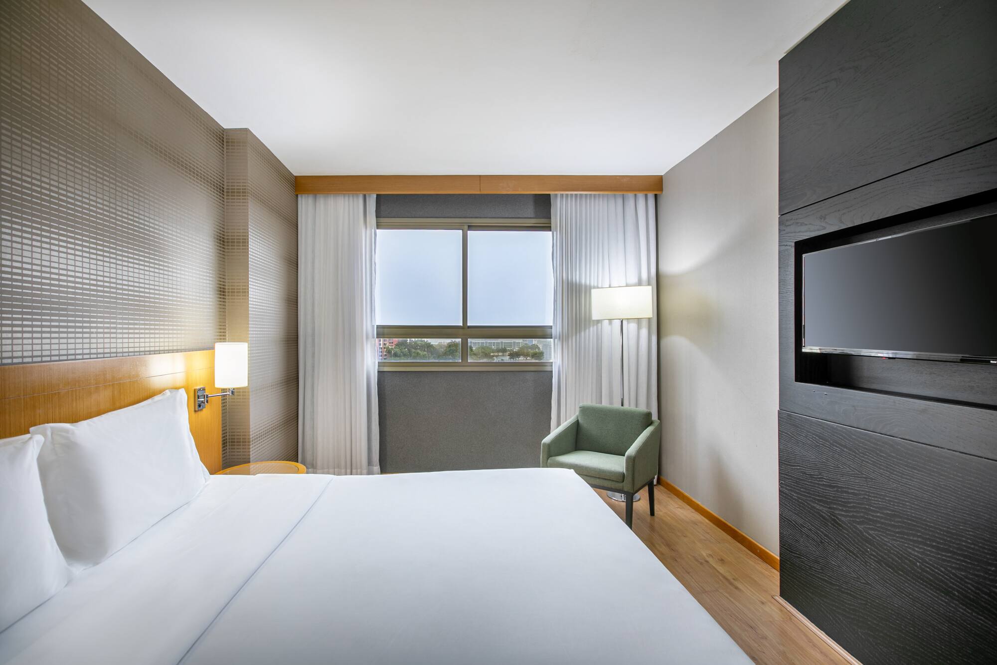 Sleek hotel room with white bed, window, armchair, and mounted flat-screen TV.