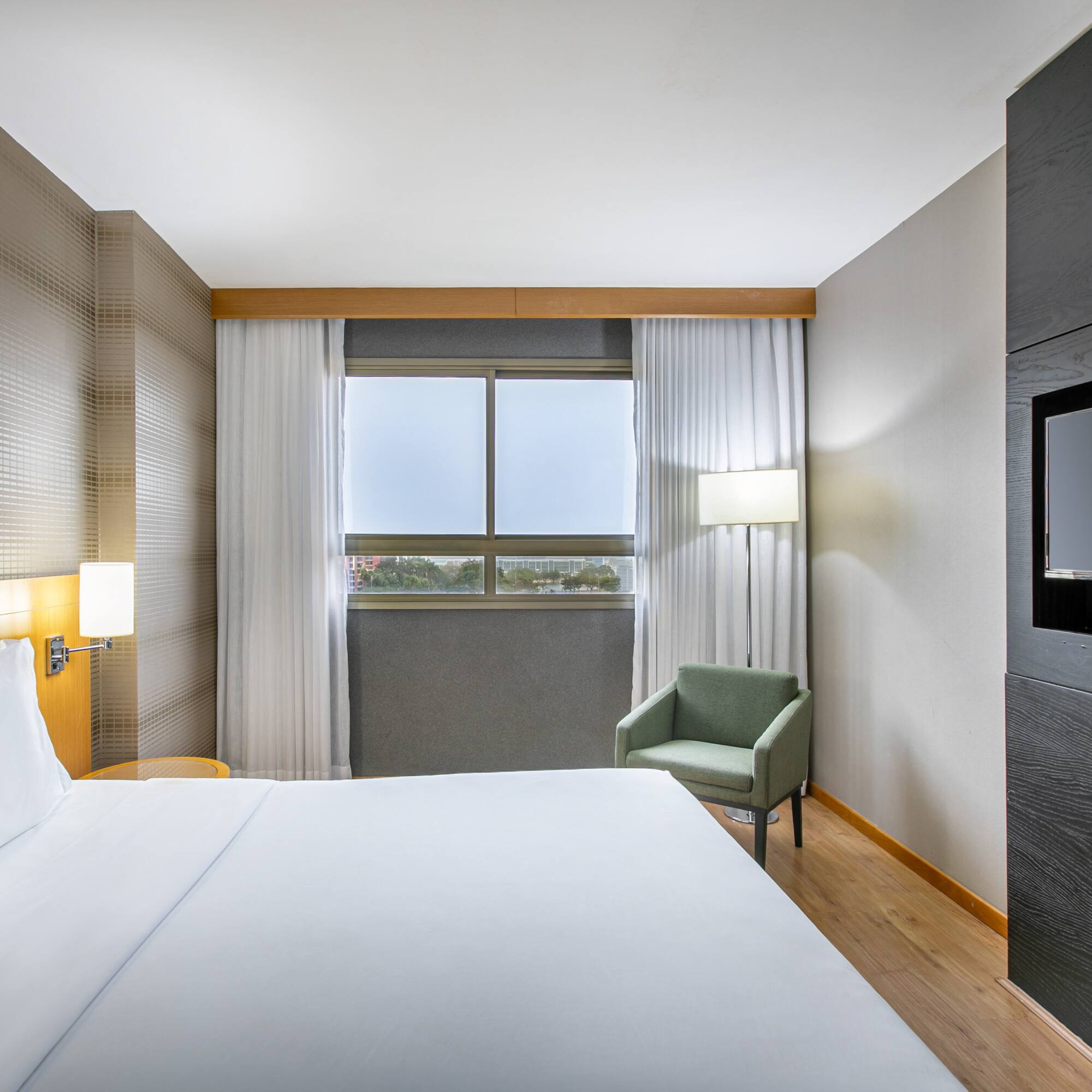 Sleek hotel room with white bed, window, armchair, and mounted flat-screen TV.