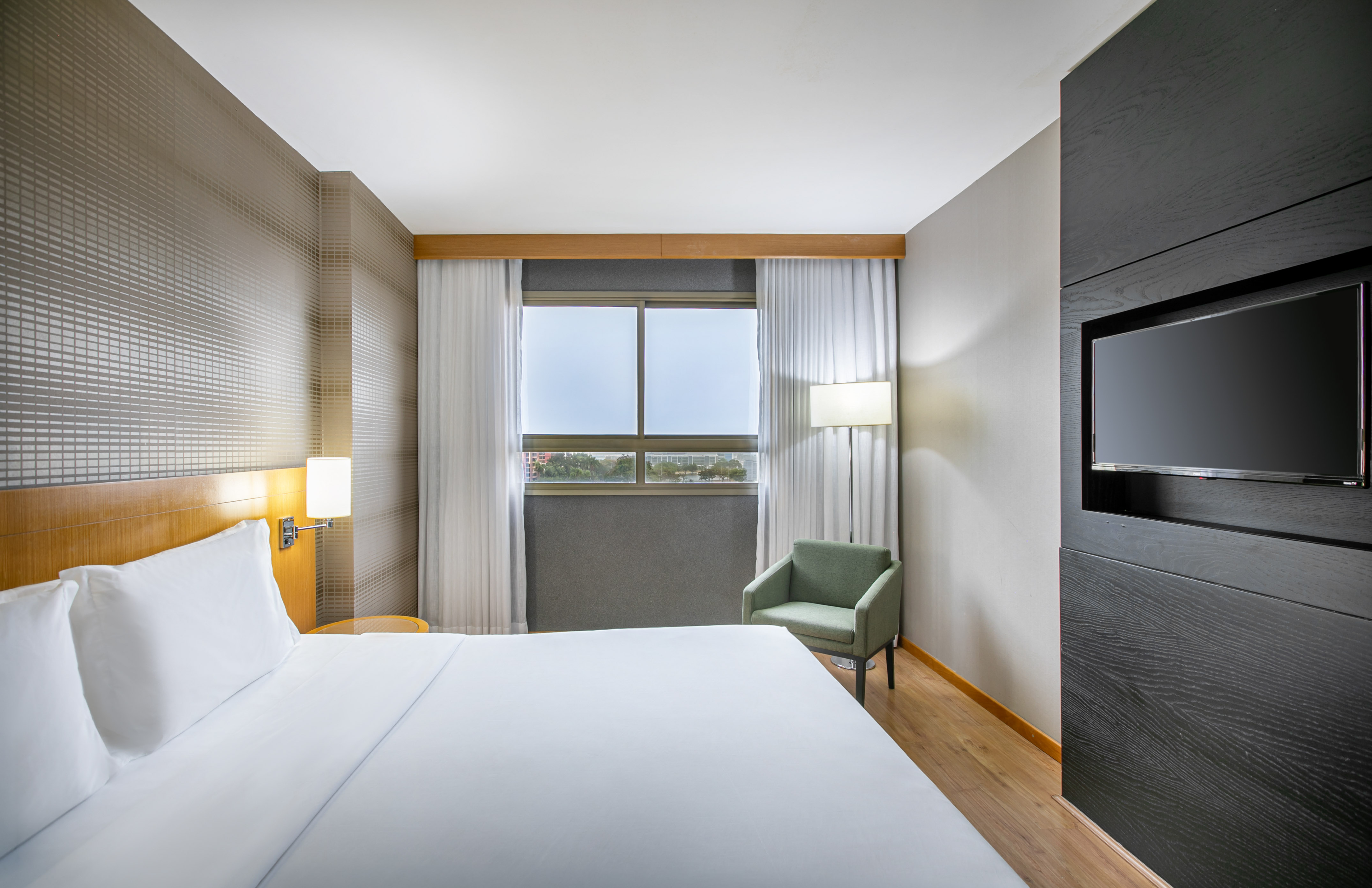 Sleek hotel room with white bed, window, armchair, and mounted flat-screen TV.