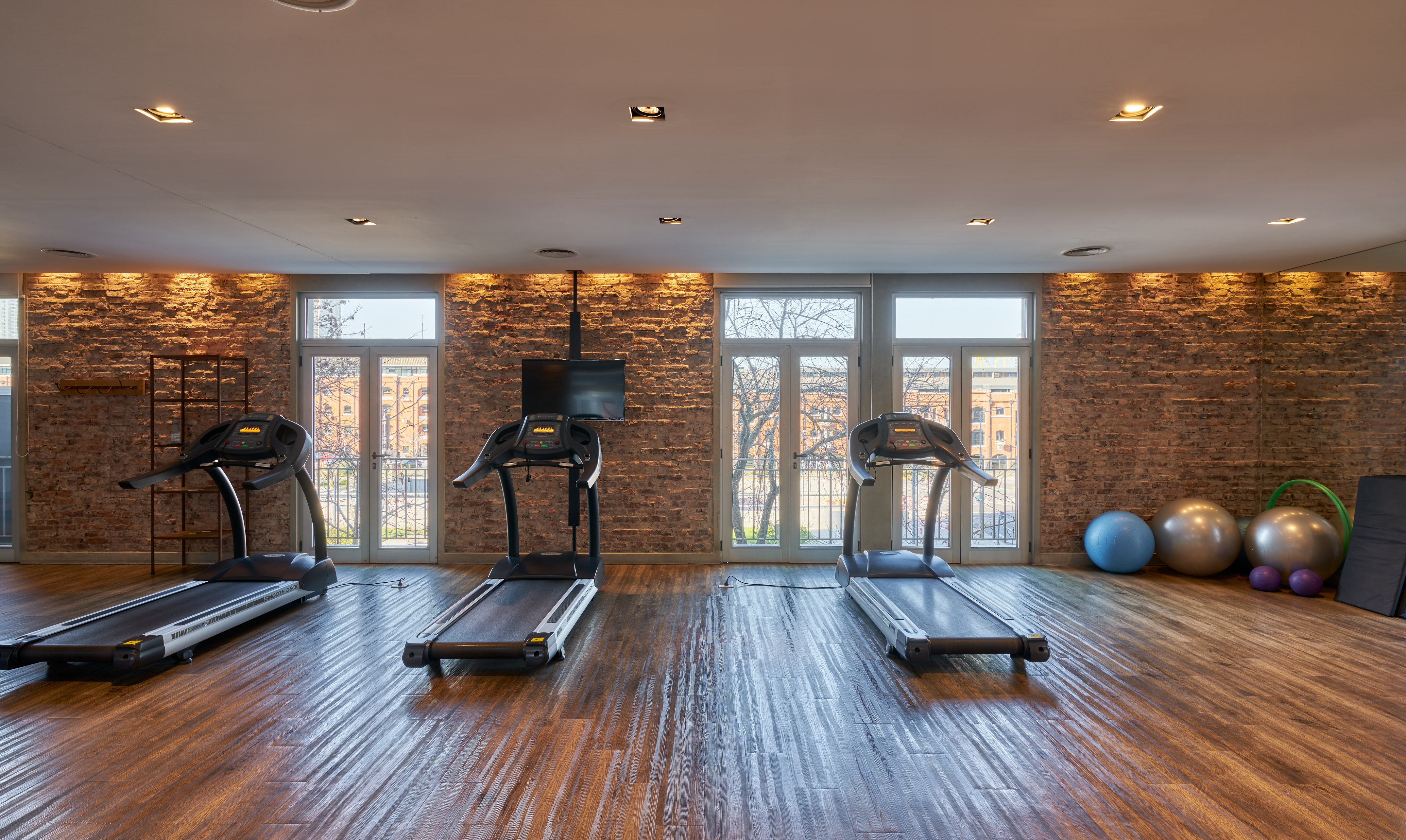 Fitness room with three treadmills, workout equipment, wood floor, and brick wall.