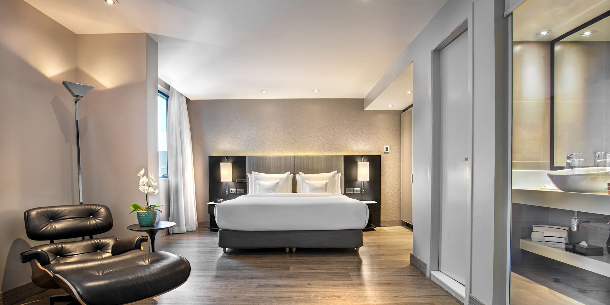 Luxury hotel bedroom suite with modern bed, Eames lounge chair, and wood floor.