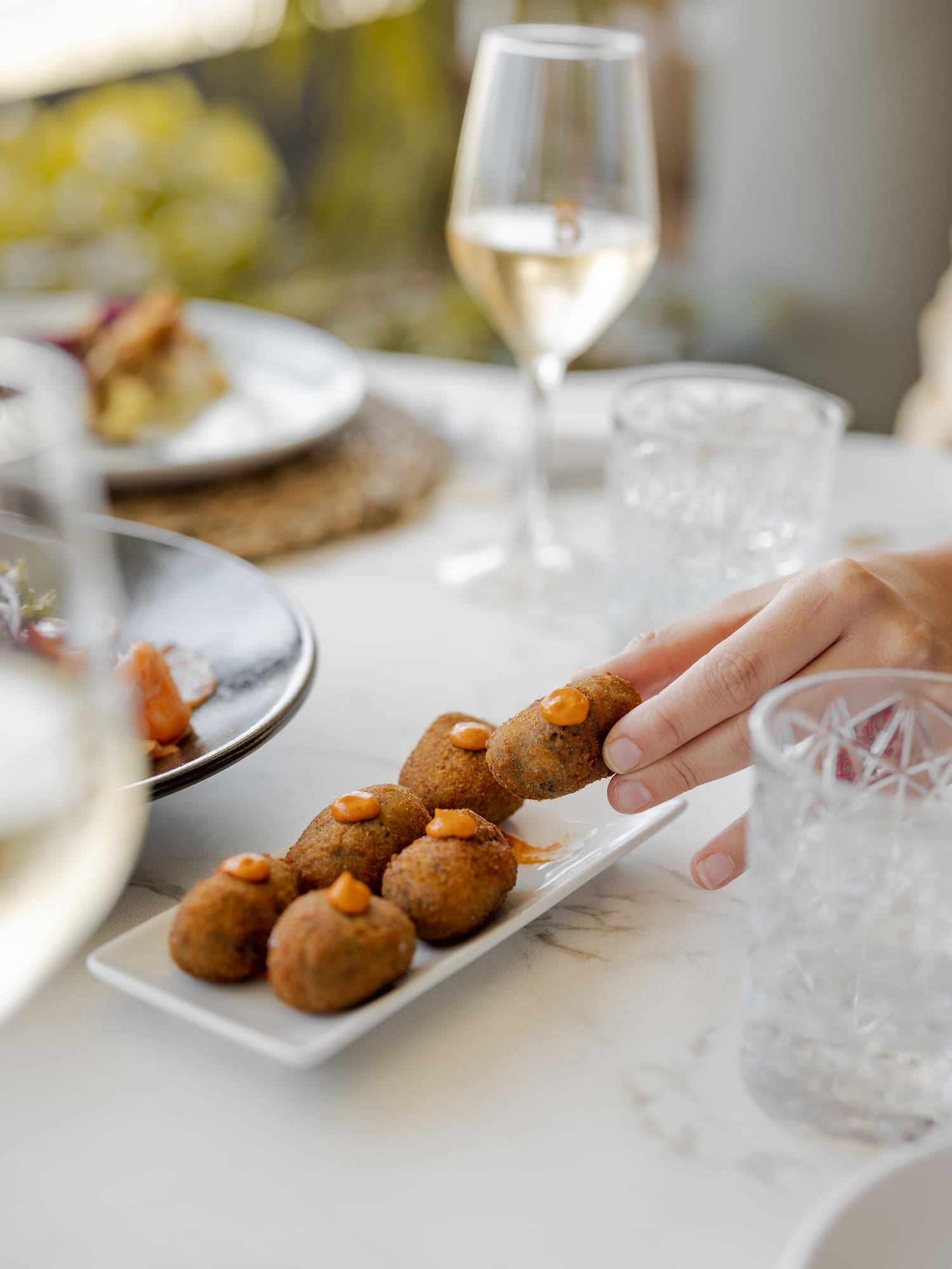 Hand picking a crispy croquette with orange sauce from a serving plate with wine.