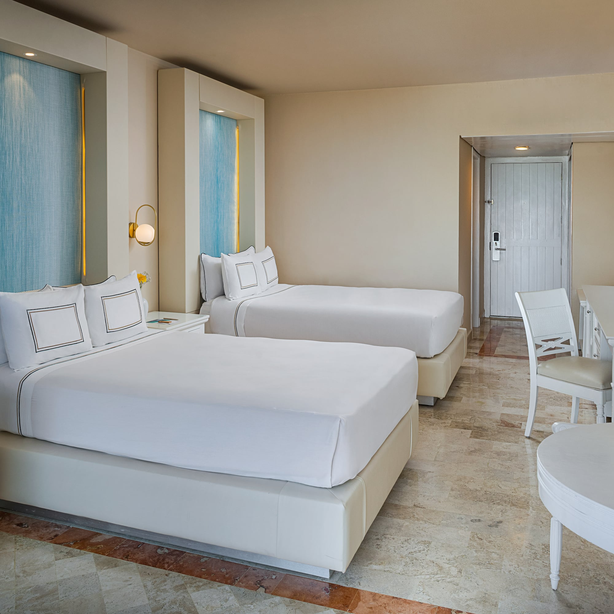 Modern hotel room with two beds, blue accents, white furniture, and tiled floor.