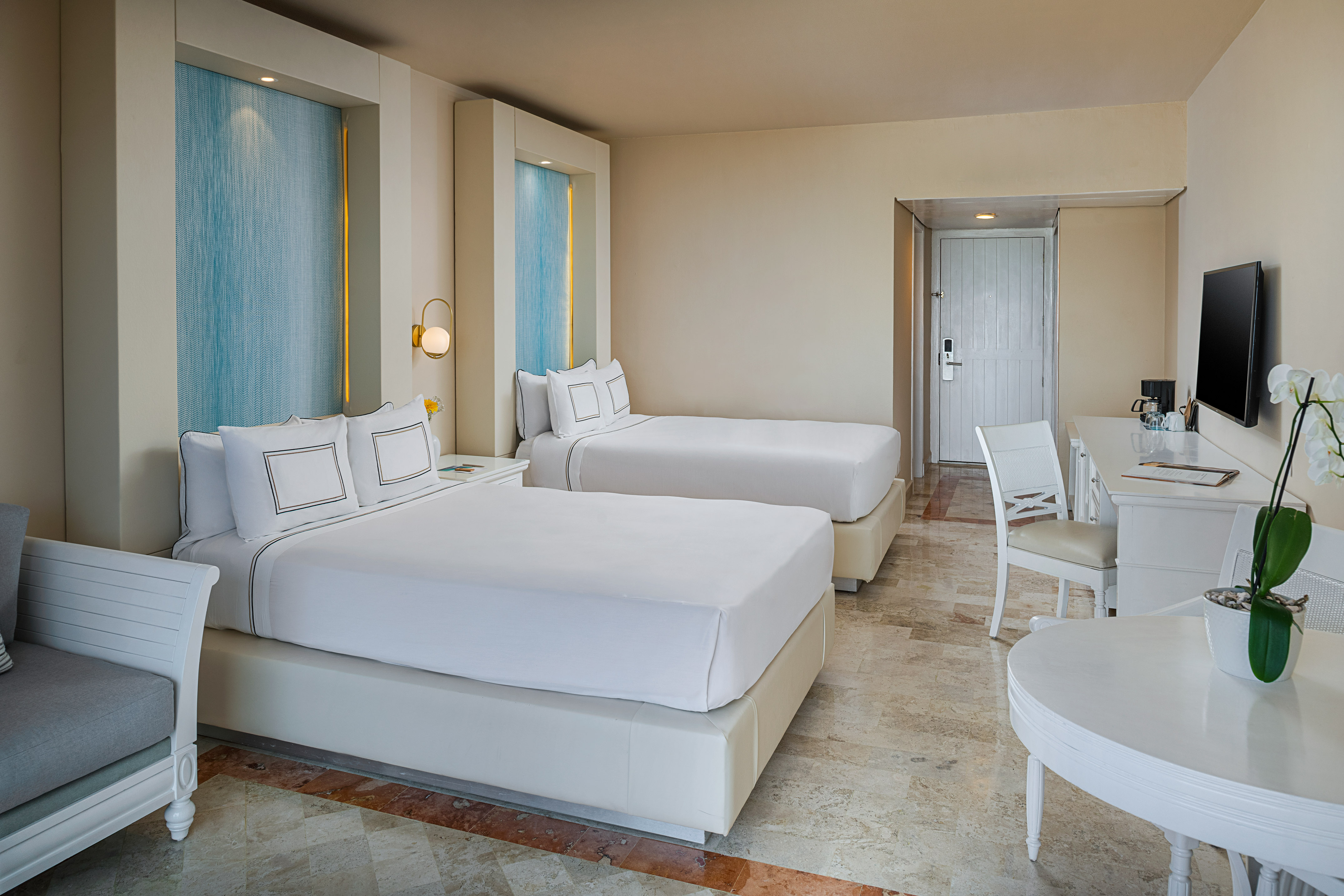 Modern hotel room with two beds, blue accents, white furniture, and tiled floor.
