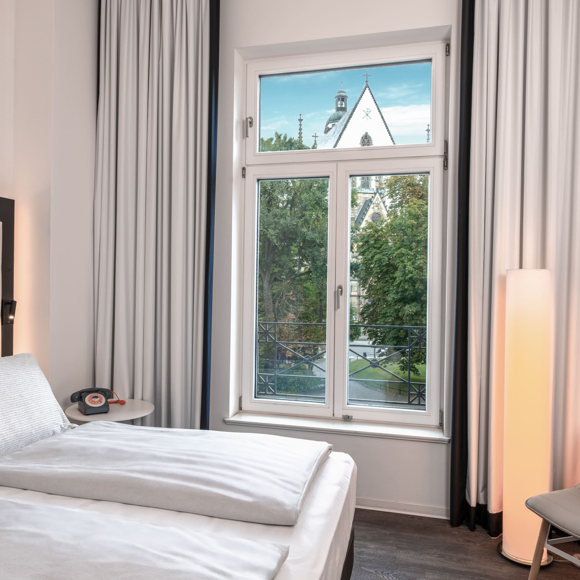 Hotel room with bed, window overlooking trees and a church steeple.