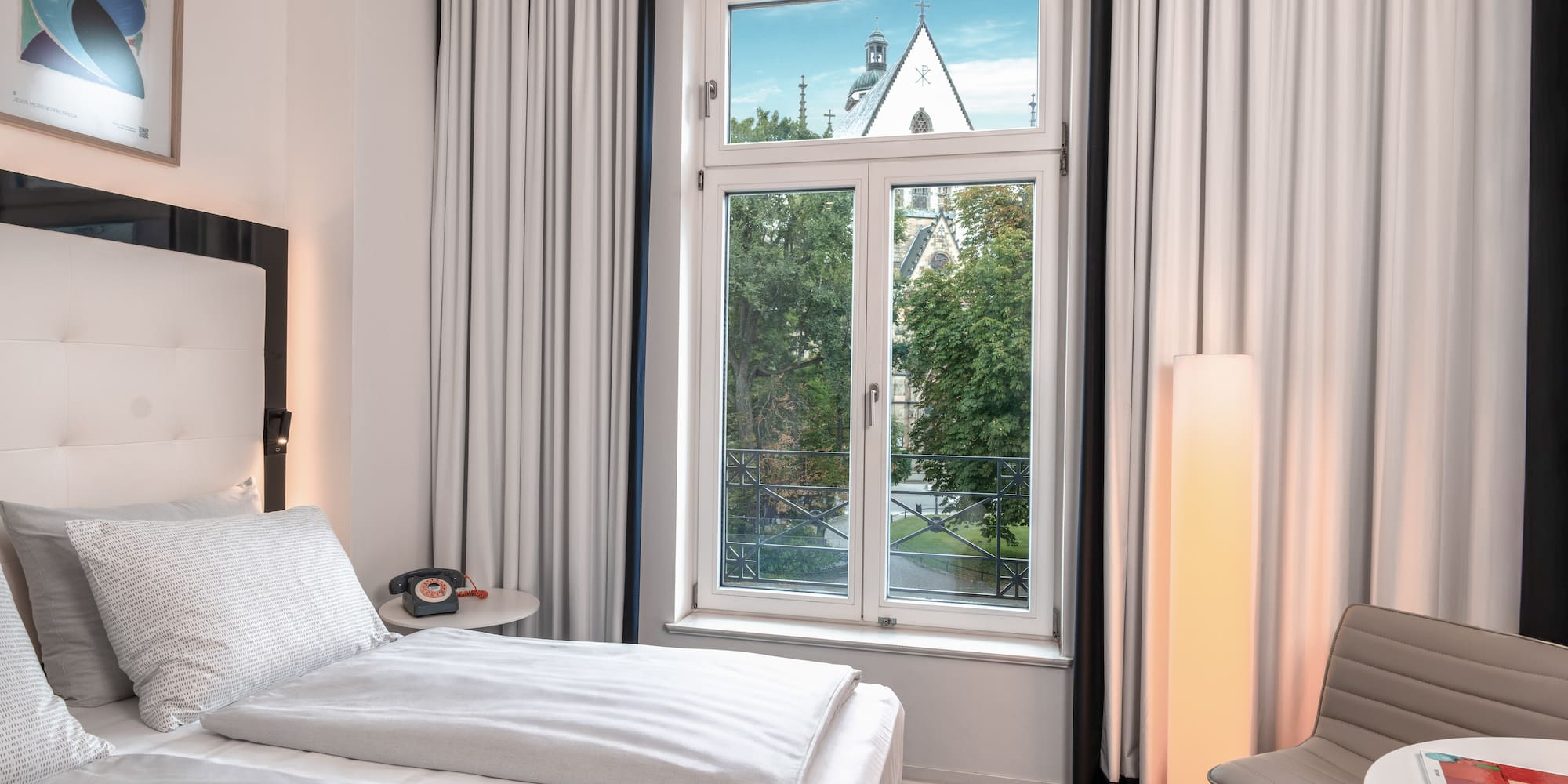 Hotel room with bed, window overlooking trees and a church steeple.