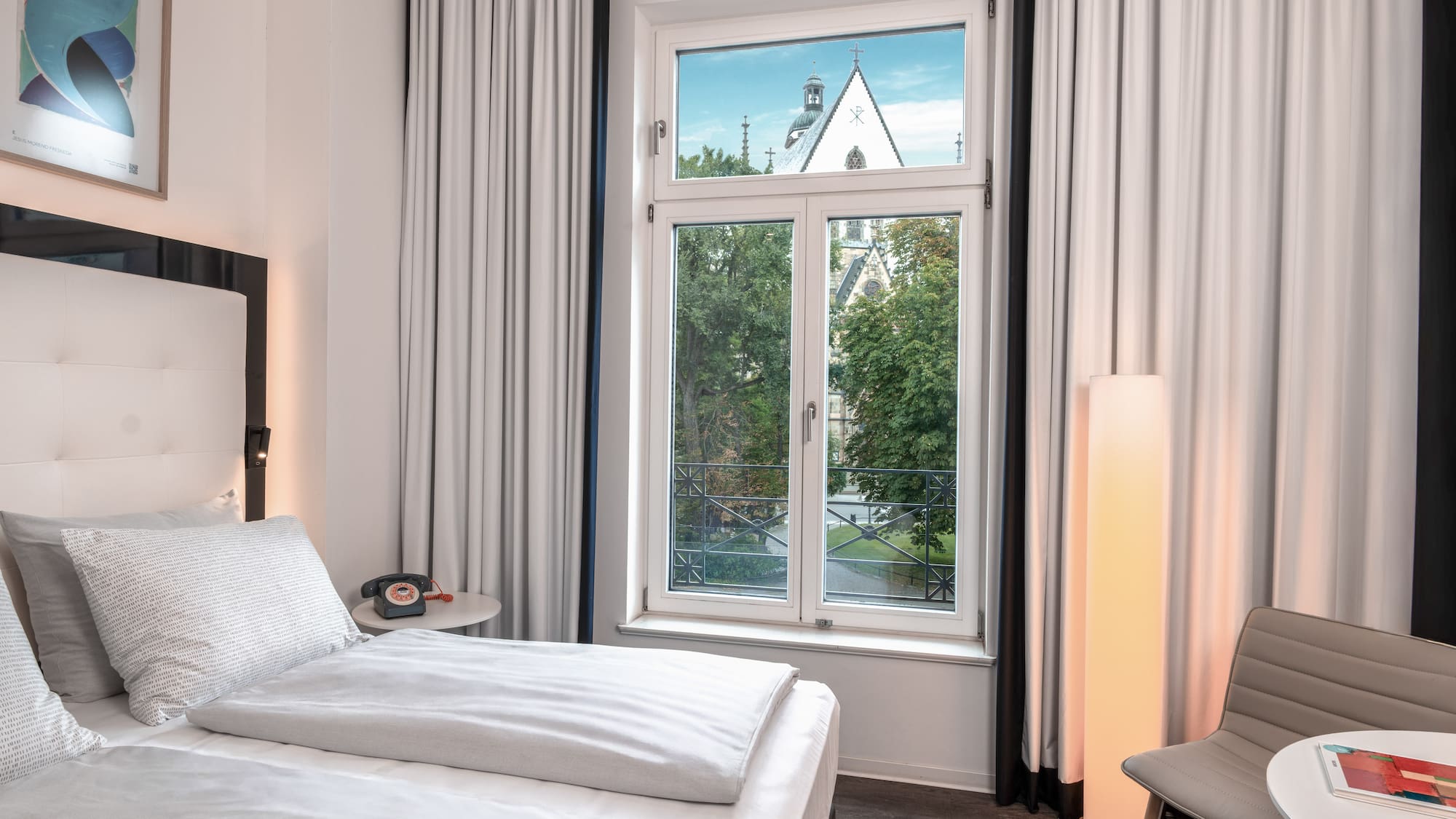 Hotel room with bed, window overlooking trees and a church steeple.
