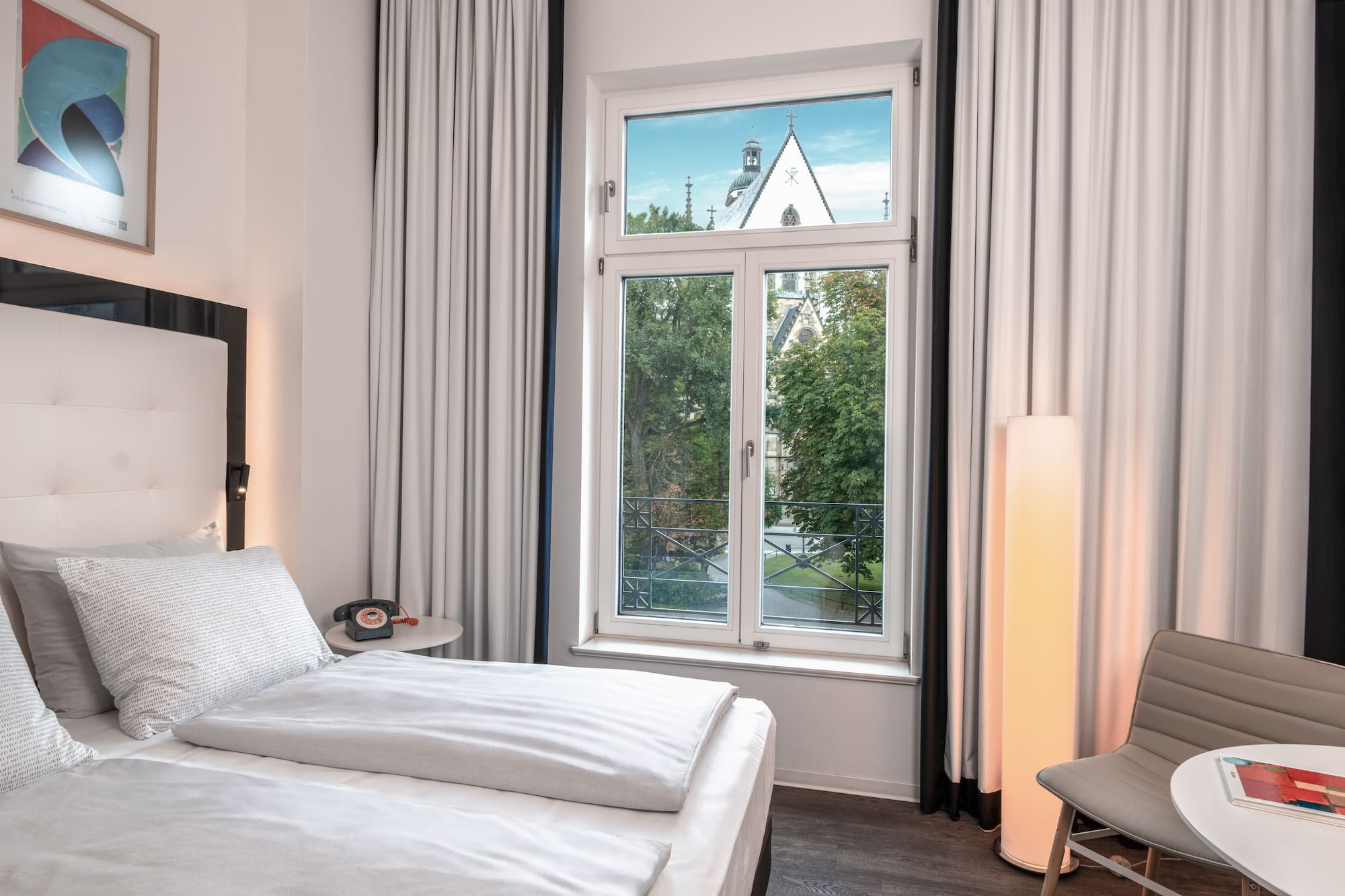 Hotel room with bed, window overlooking trees and a church steeple.