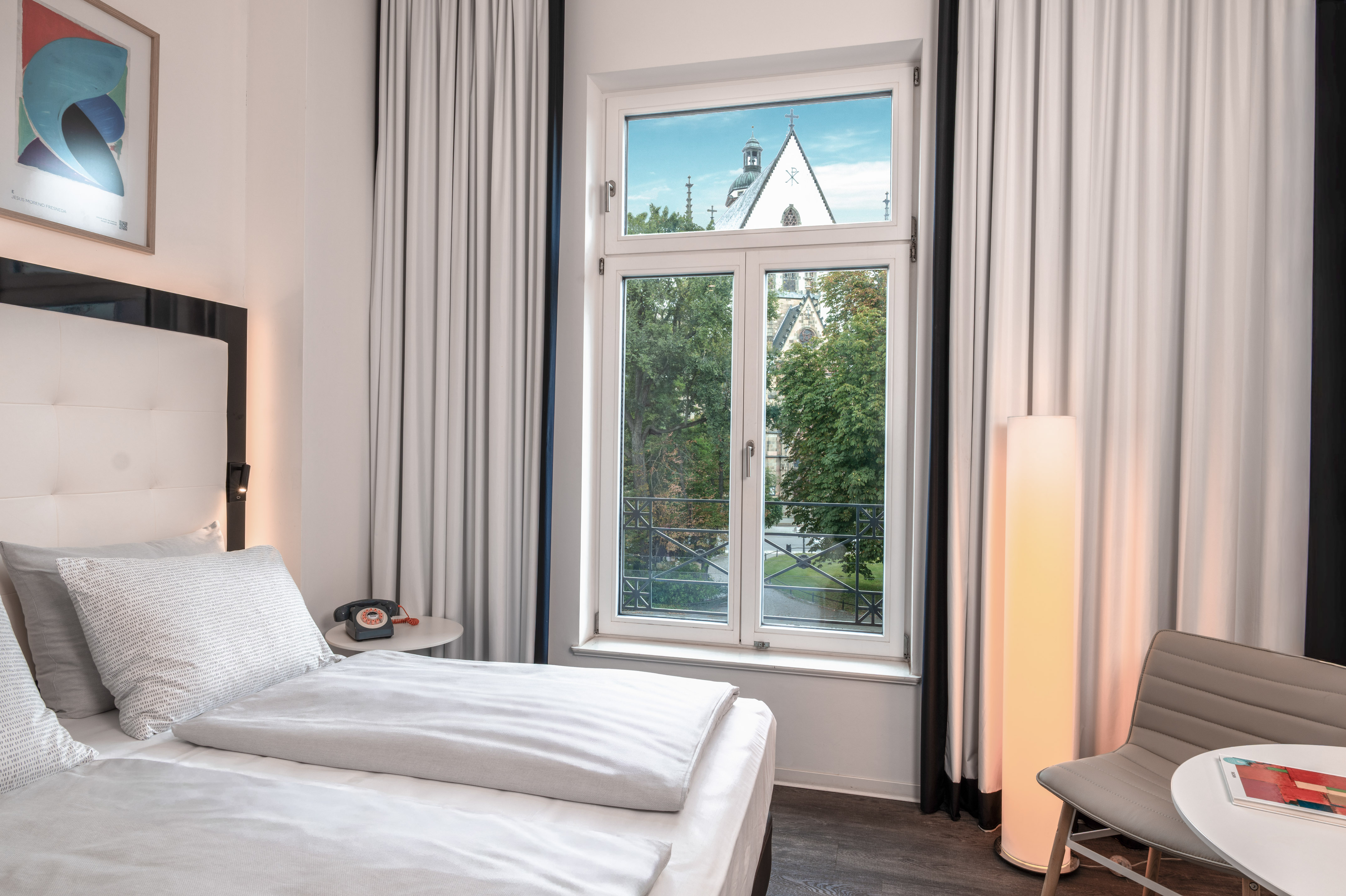 Hotel room with bed, window overlooking trees and a church steeple.