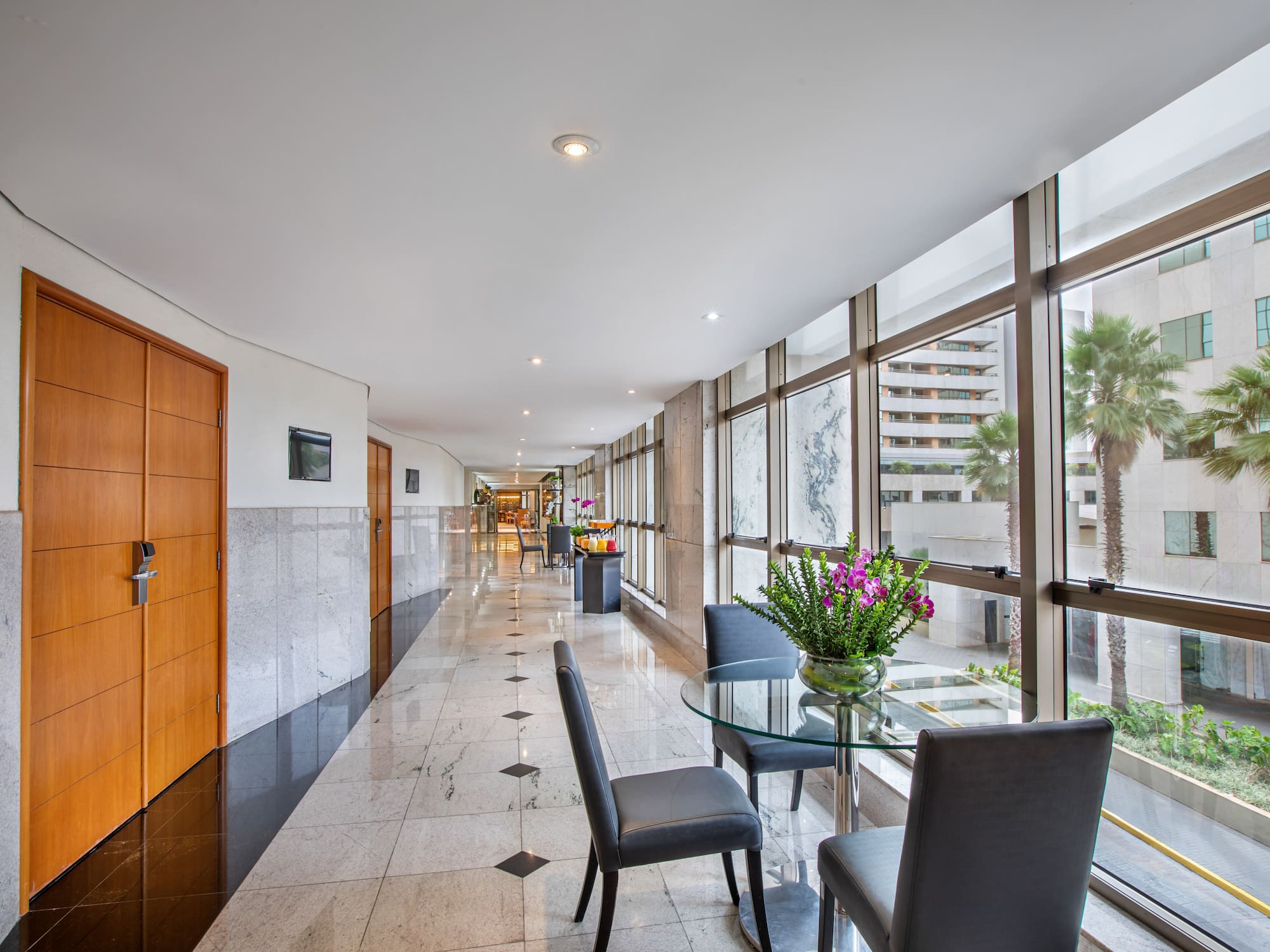 Modern hotel corridor with marble floor, window seating, and palm trees.