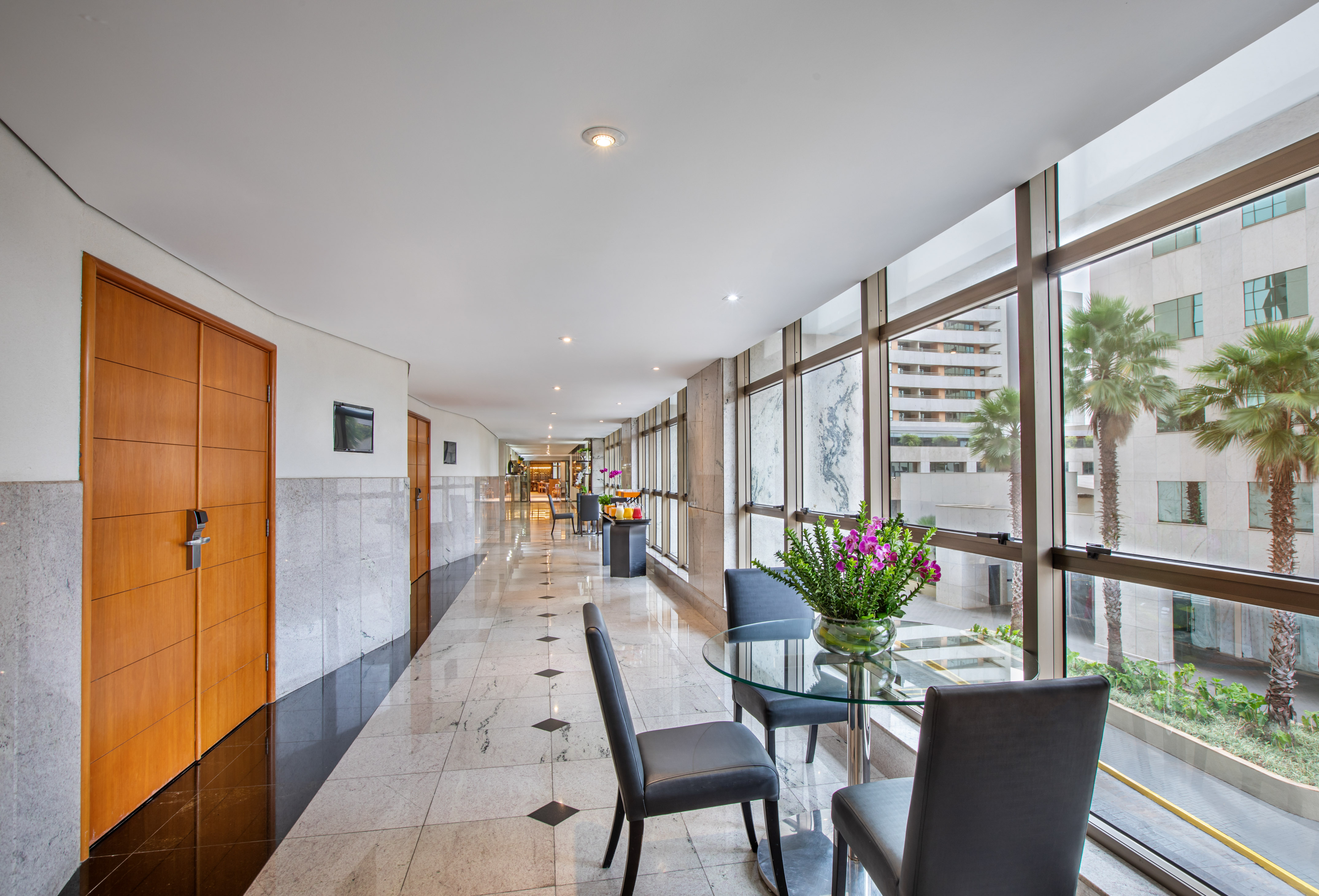 Modern hotel corridor with marble floor, window seating, and palm trees.