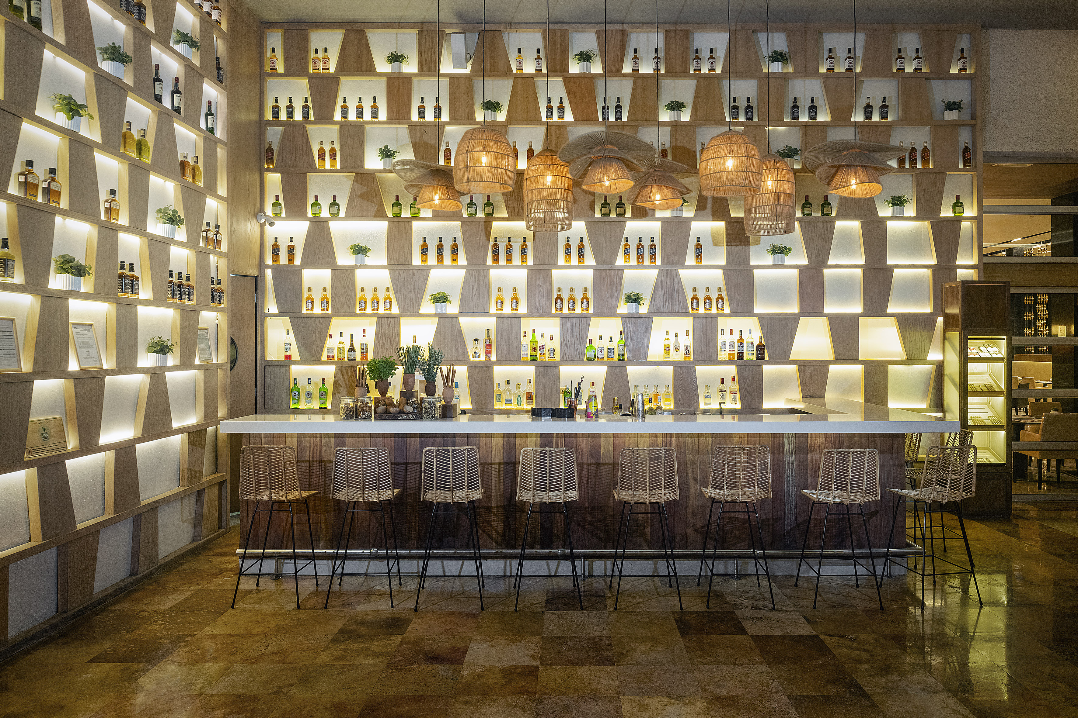 Well-lit modern bar with extensive liquor bottle display and rattan barstools.