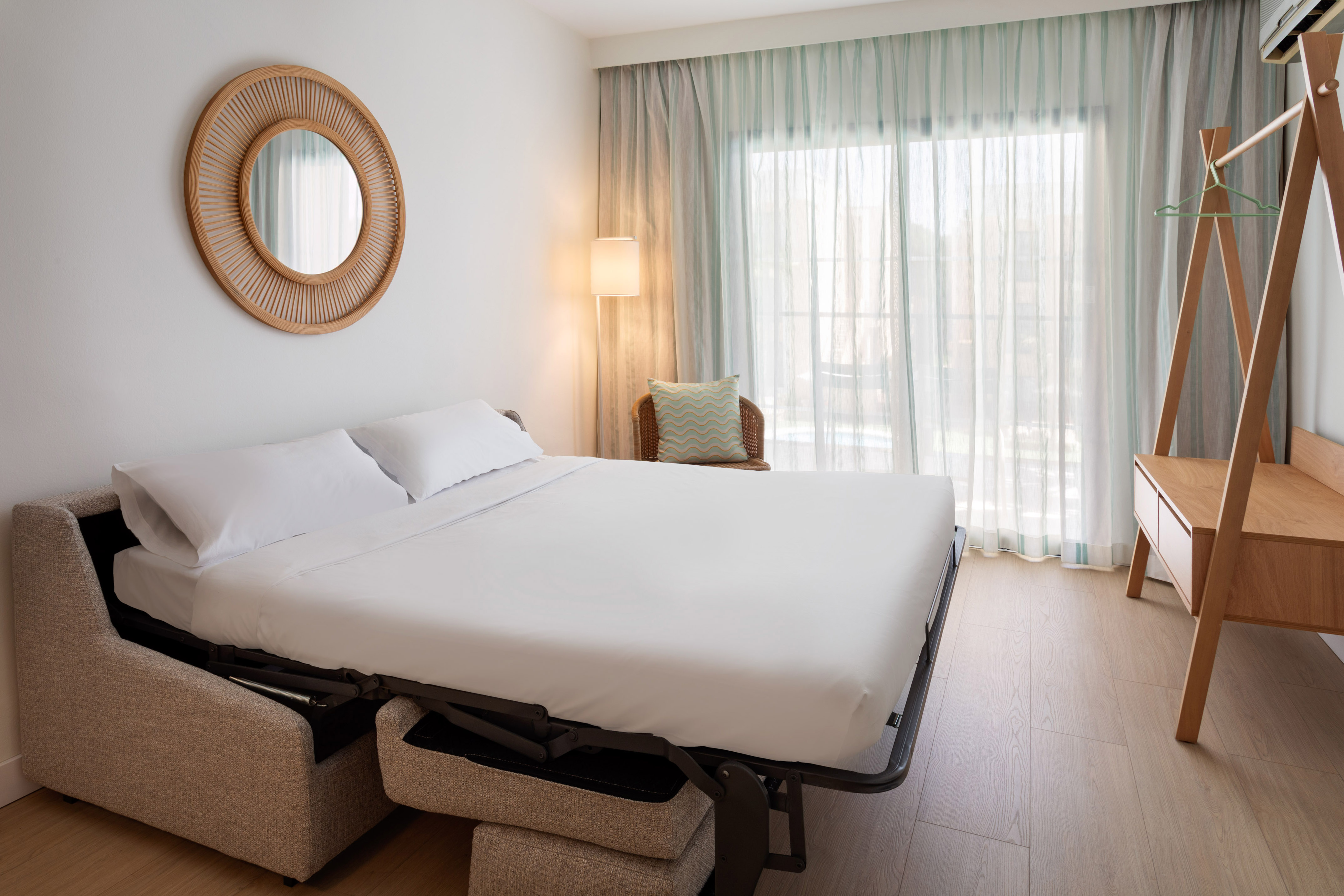 Bright hotel room with modern pull-out sofa bed and round rattan mirror.