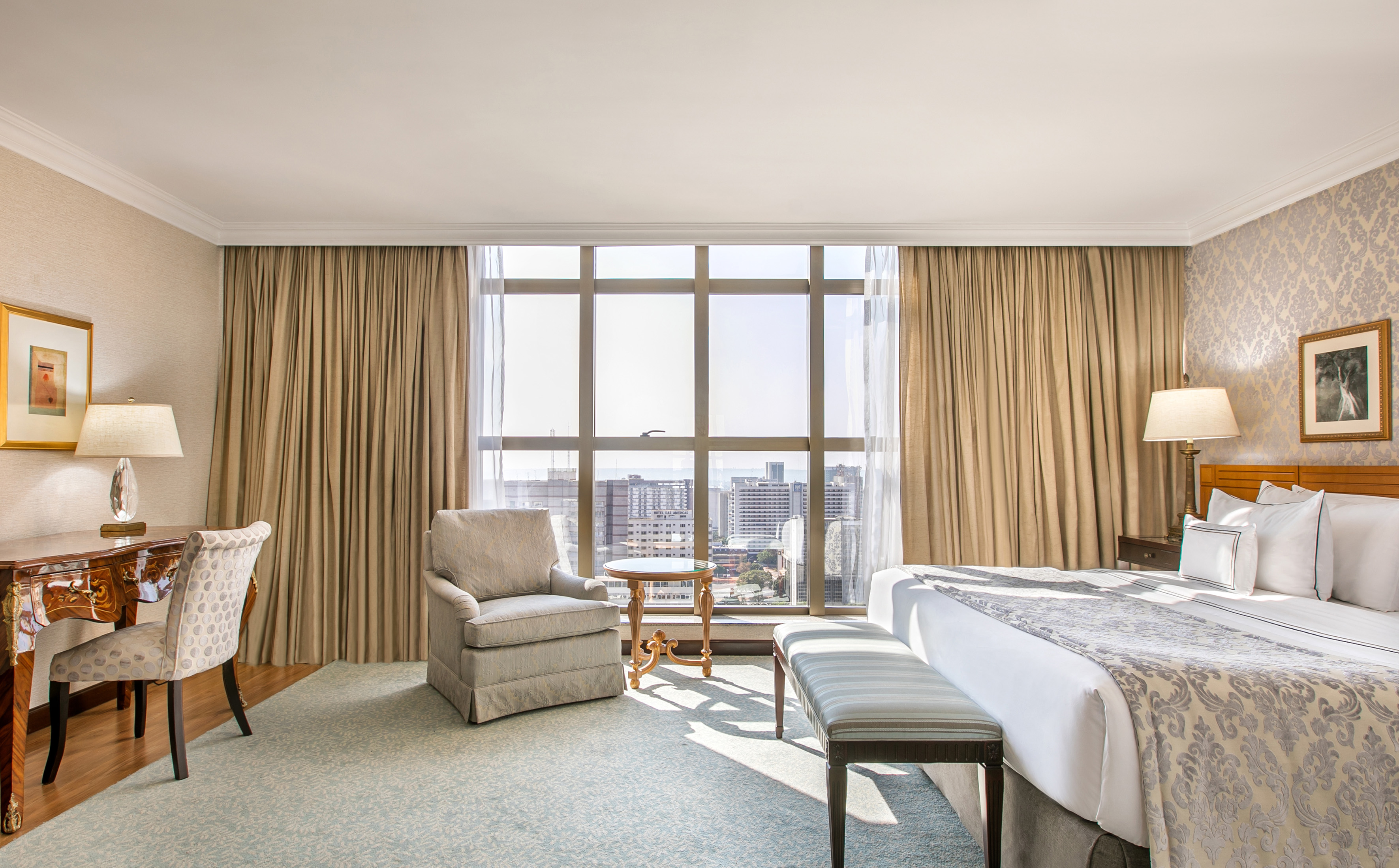 Elegant hotel room with king bed, desk, armchair, and city skyline view.