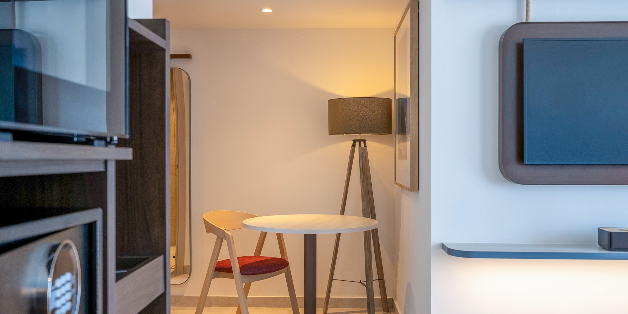 Hotel room interior: safe, small table with chair, floor lamp, wall TV.