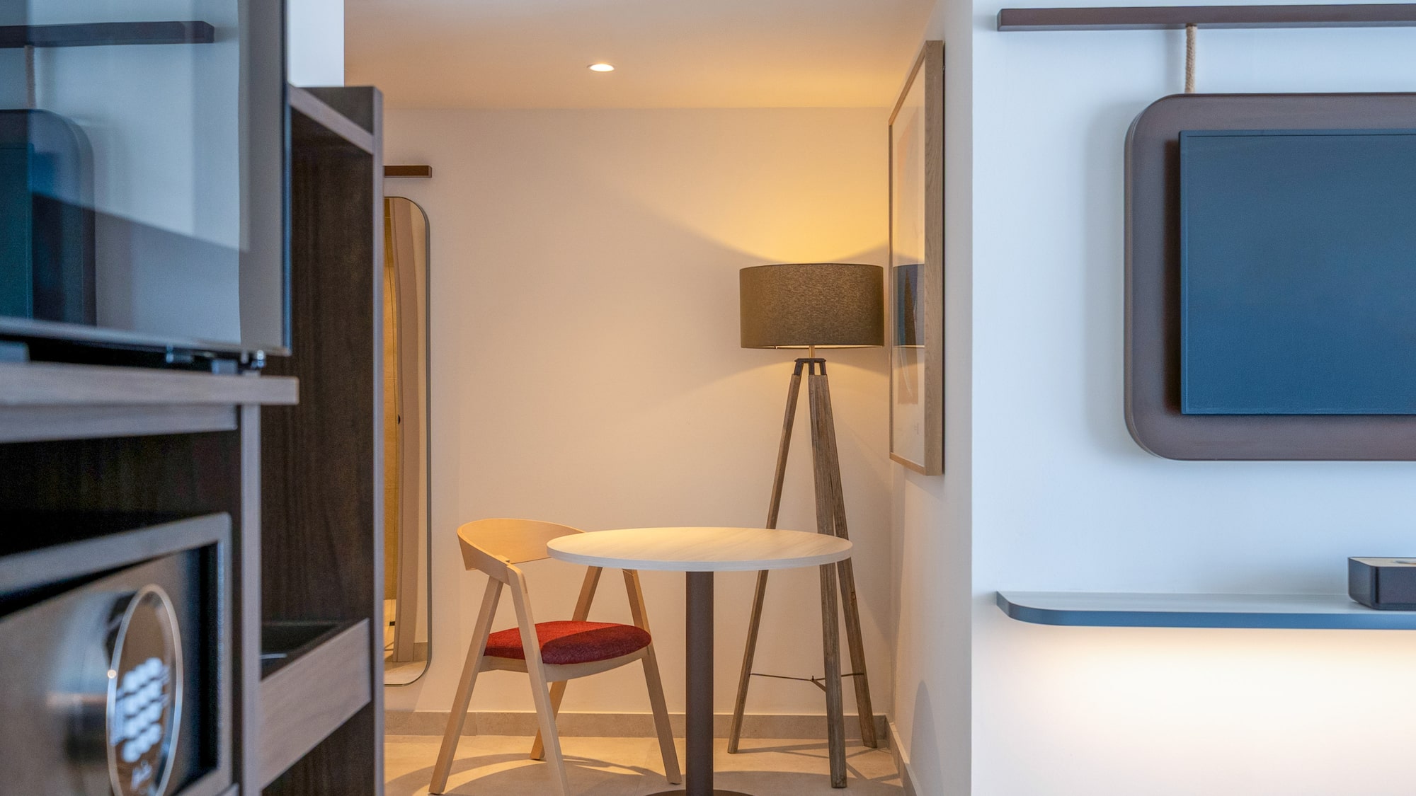 Hotel room interior: safe, small table with chair, floor lamp, wall TV.