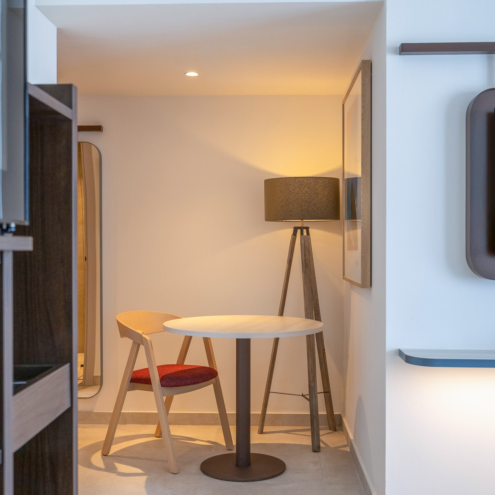 Hotel room interior: safe, small table with chair, floor lamp, wall TV.