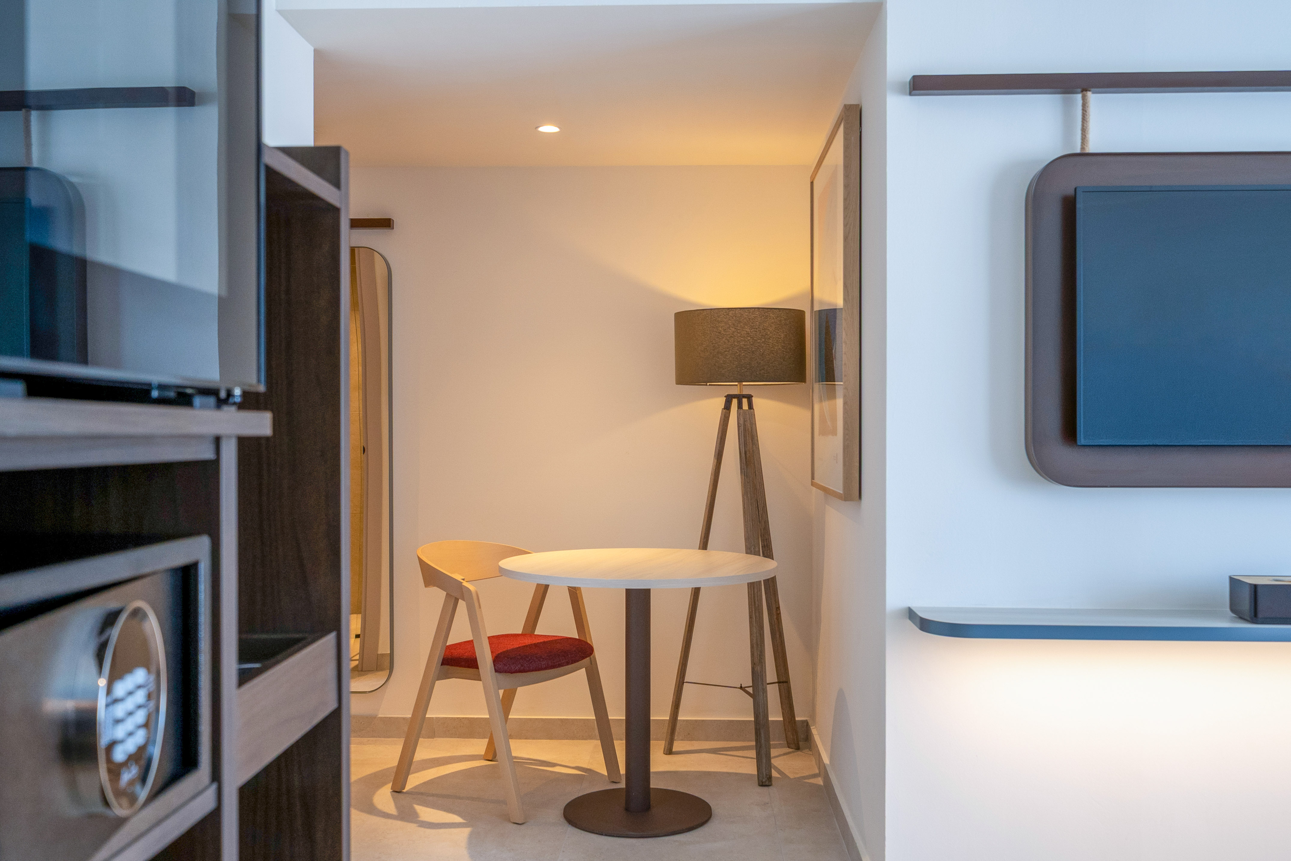 Hotel room interior: safe, small table with chair, floor lamp, wall TV.