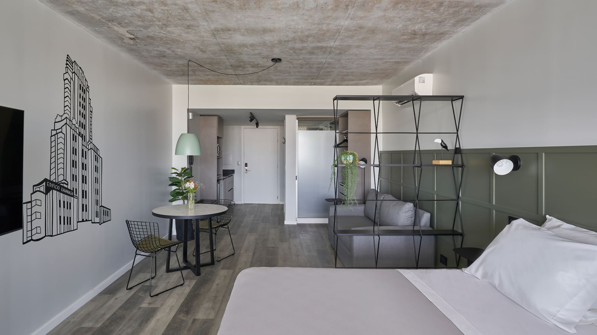 Modern studio apartment with grey bed, concrete ceiling, green wall, and dining area.