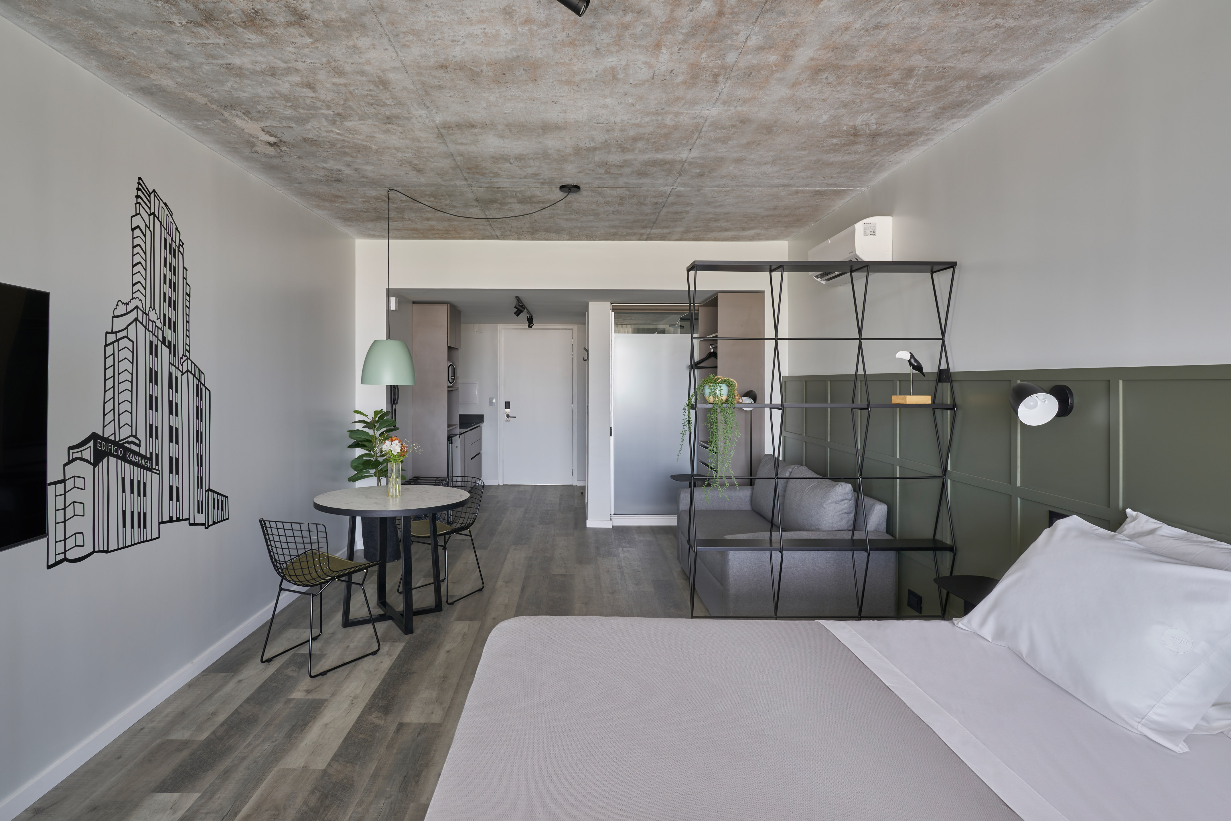 Modern studio apartment with grey bed, concrete ceiling, green wall, and dining area.