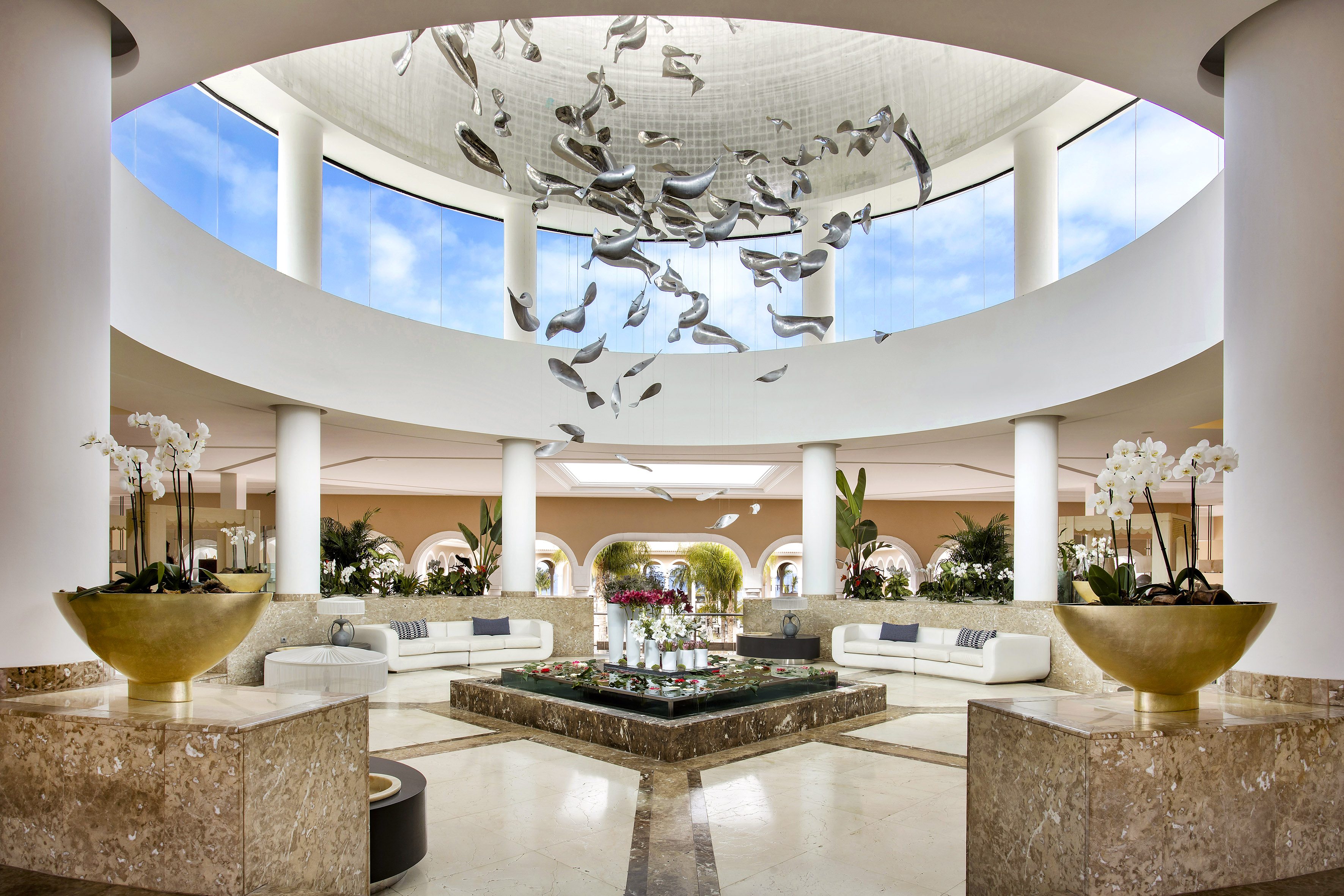 Grand hotel lobby with fish sculpture chandelier, central fountain, and orchids.