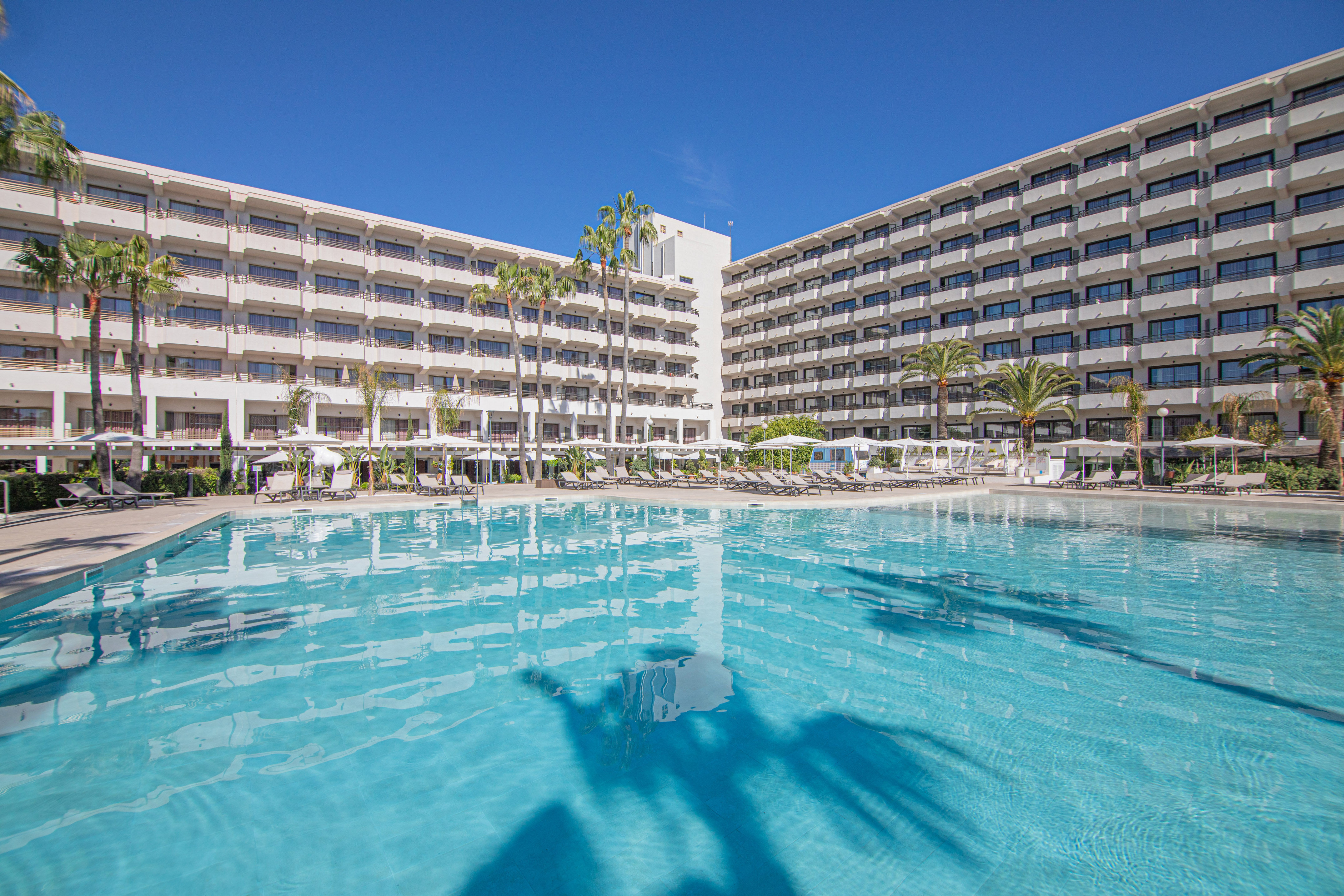 Sunny resort hotel with large swimming pool, palm trees, and lounge area.
