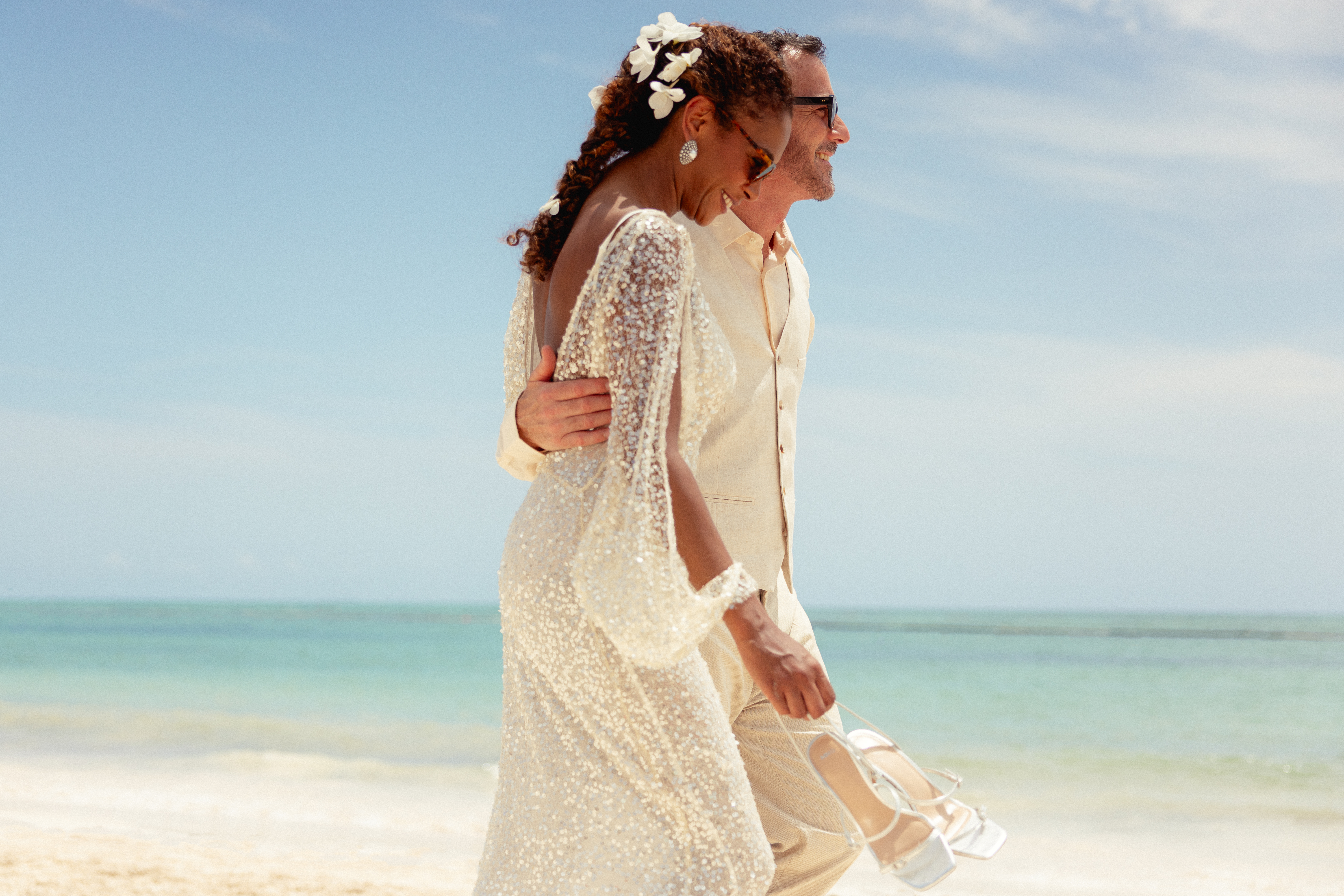 Wedding couple in sequined dress, linen suit, walking on tropical beach.