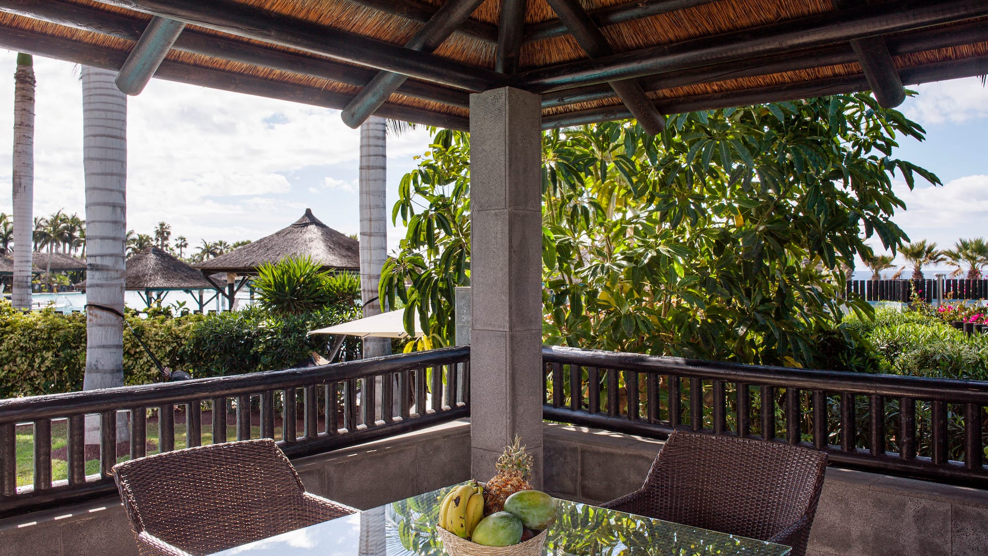 Terraza cubierta con mesa y frutas, techo de paja, palmeras tropicales al fondo.