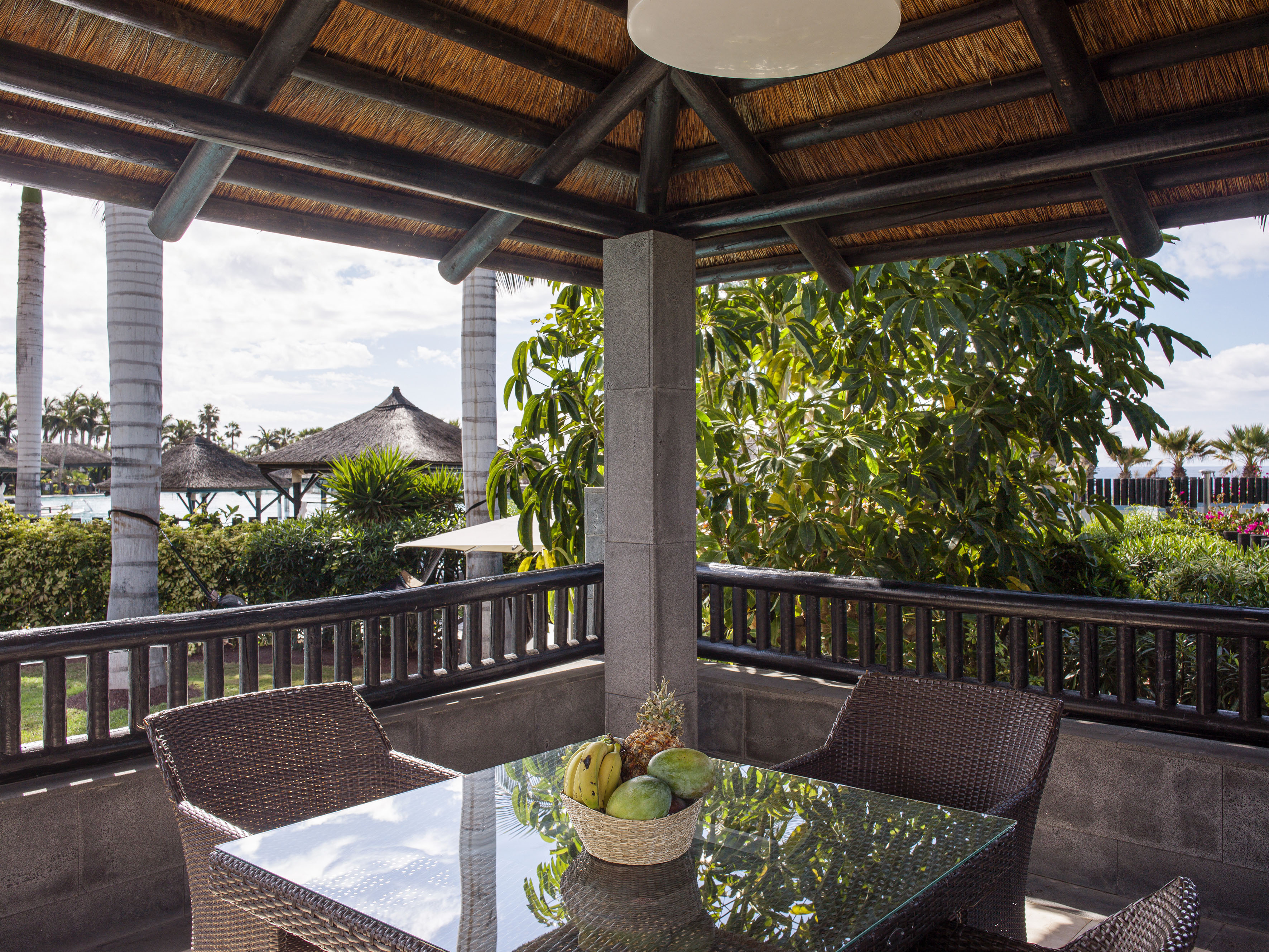 Terraza cubierta con mesa y frutas, techo de paja, palmeras tropicales al fondo.