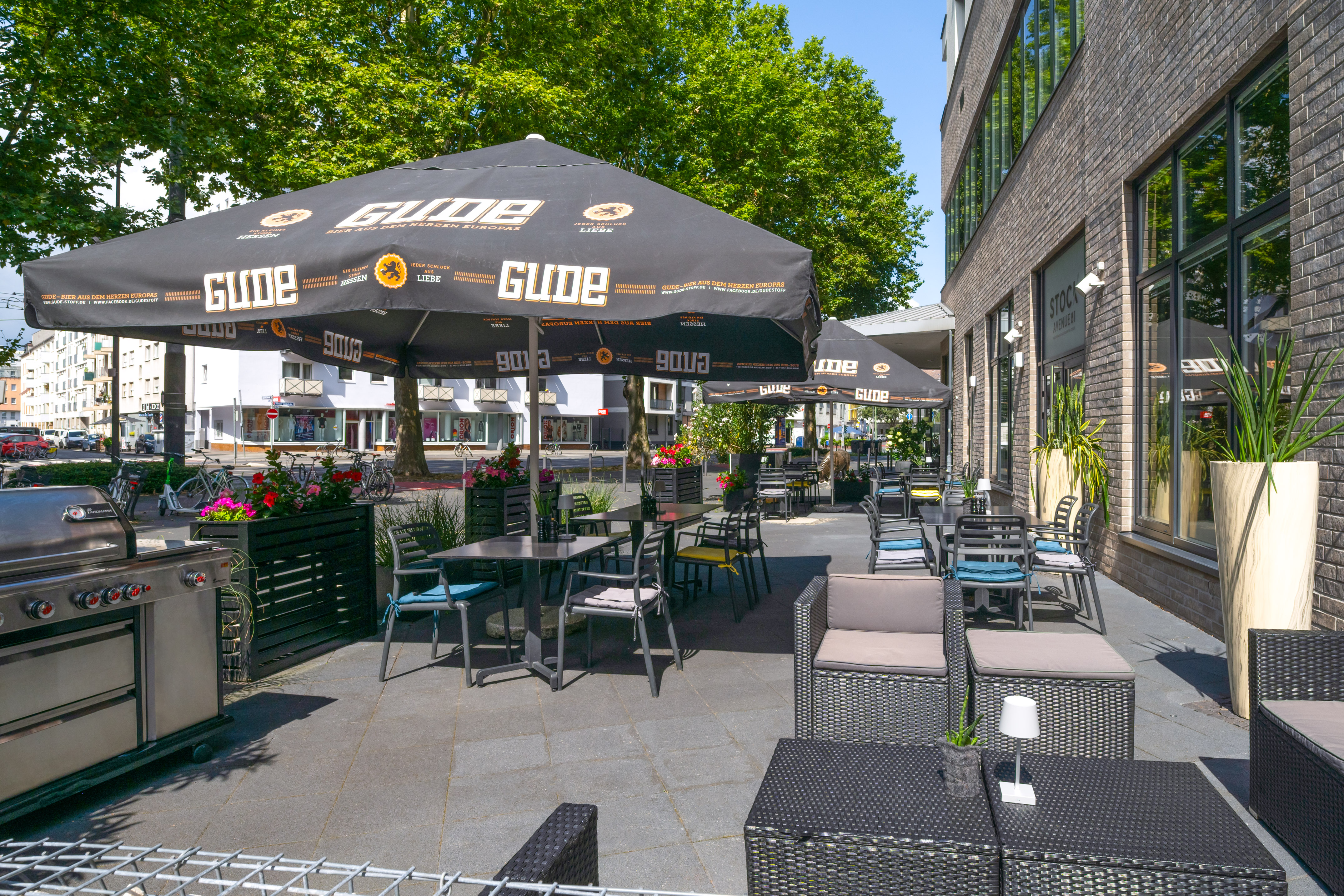 Outdoor GUDE beer garden patio with tables, chairs, umbrellas, and BBQ grill.