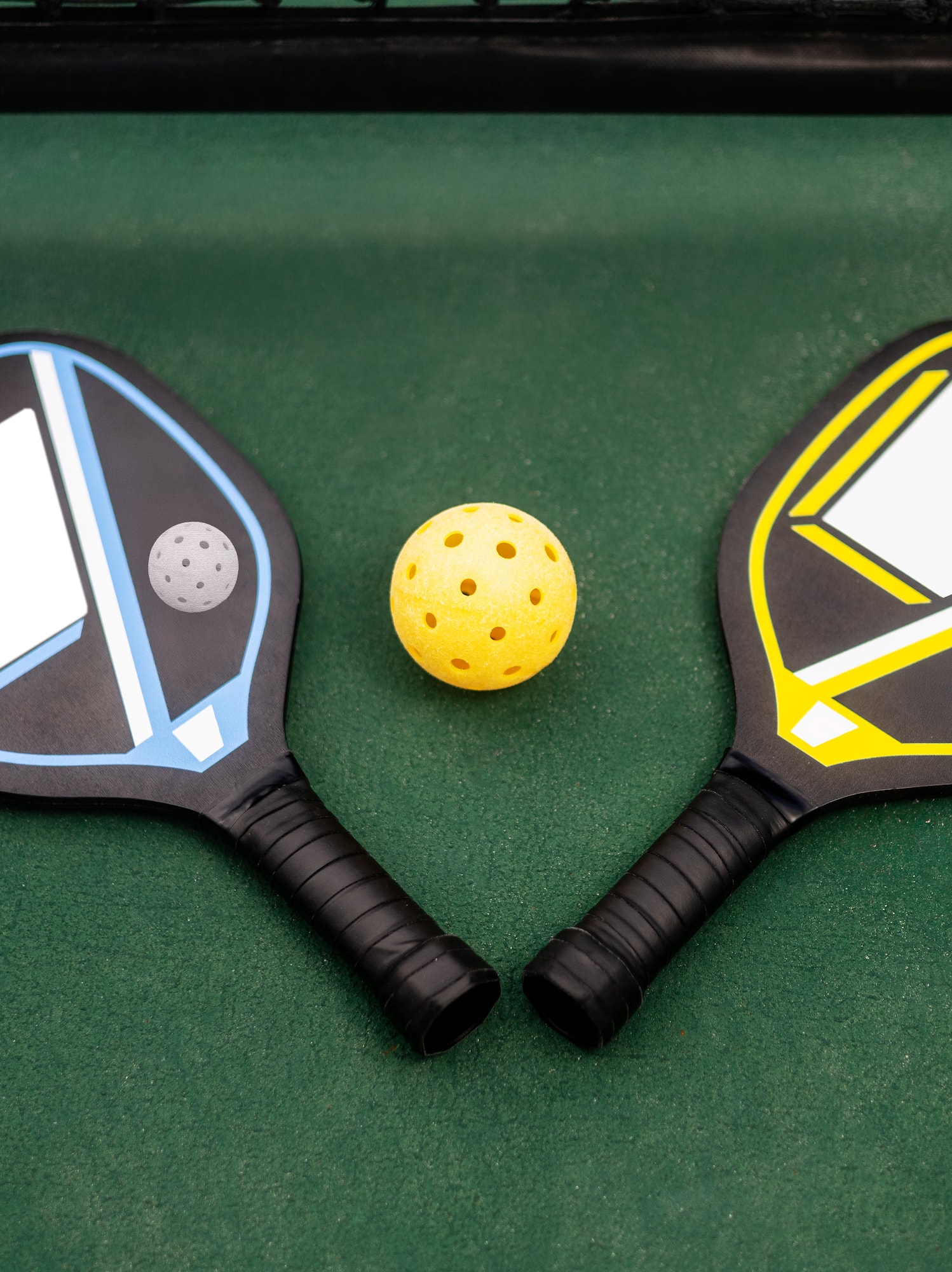 Palas de pickleball azul y amarilla, pelota en cancha verde.