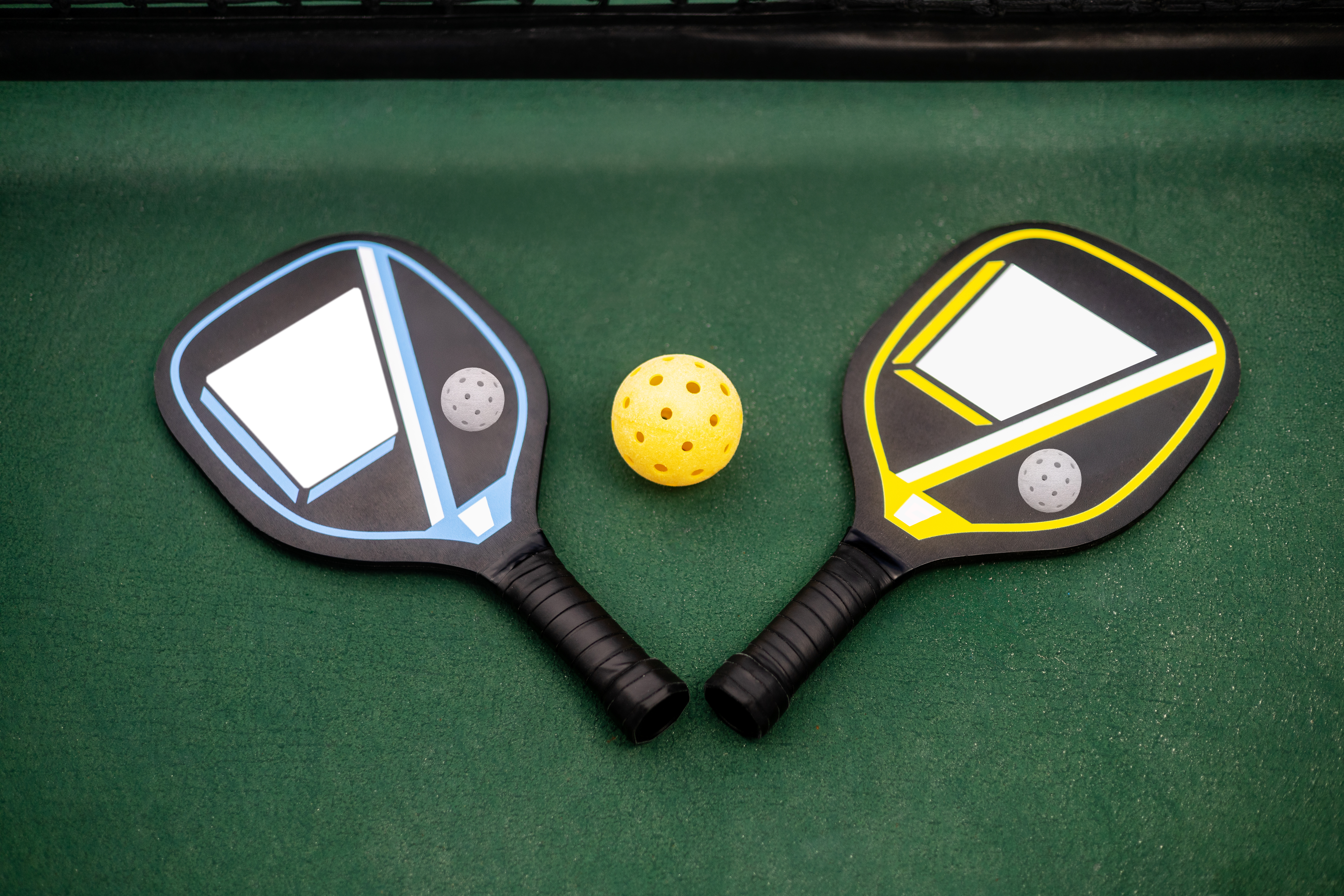Palas de pickleball azul y amarilla, pelota en cancha verde.