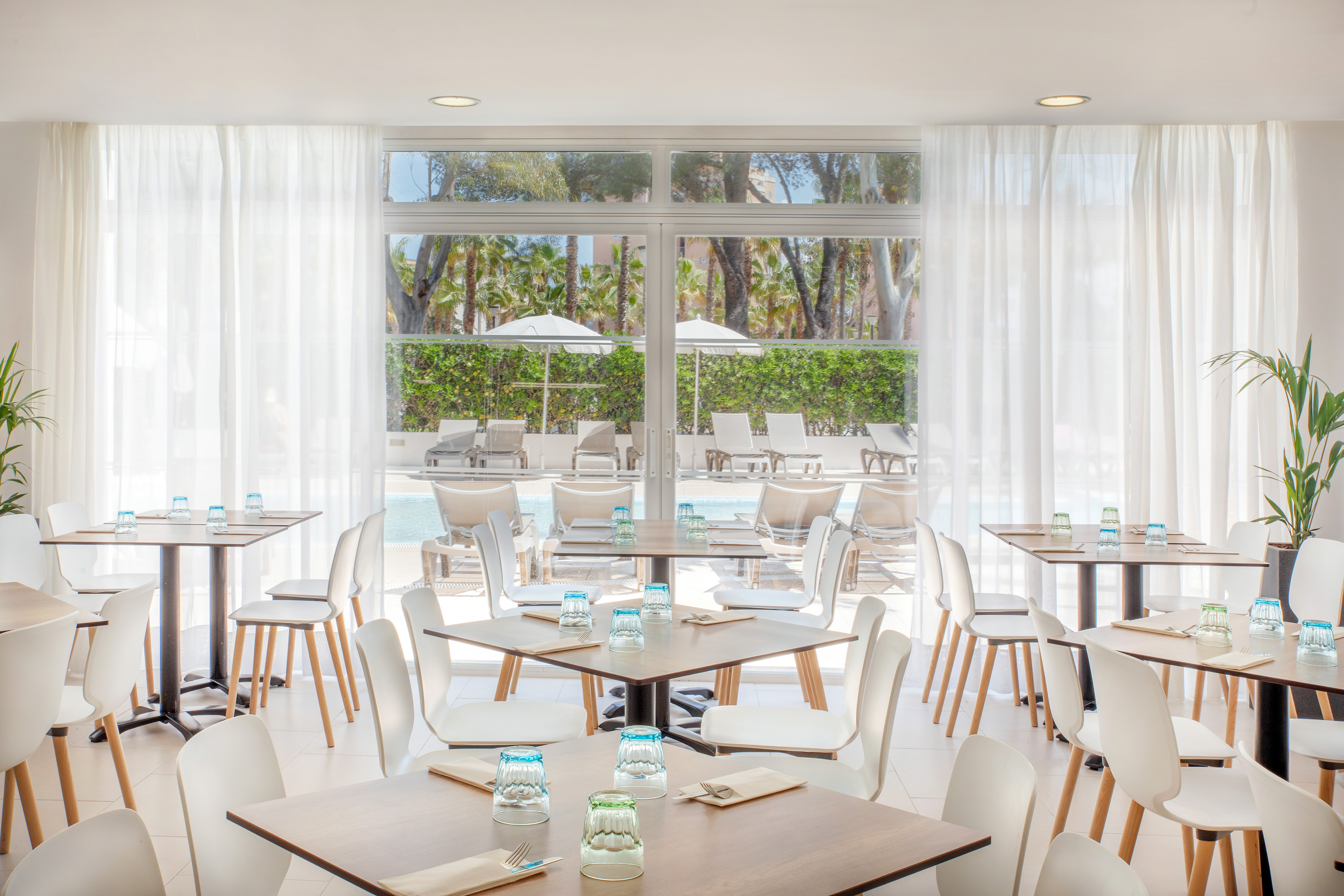 Bright, modern restaurant with poolside view, white chairs, and palm trees.