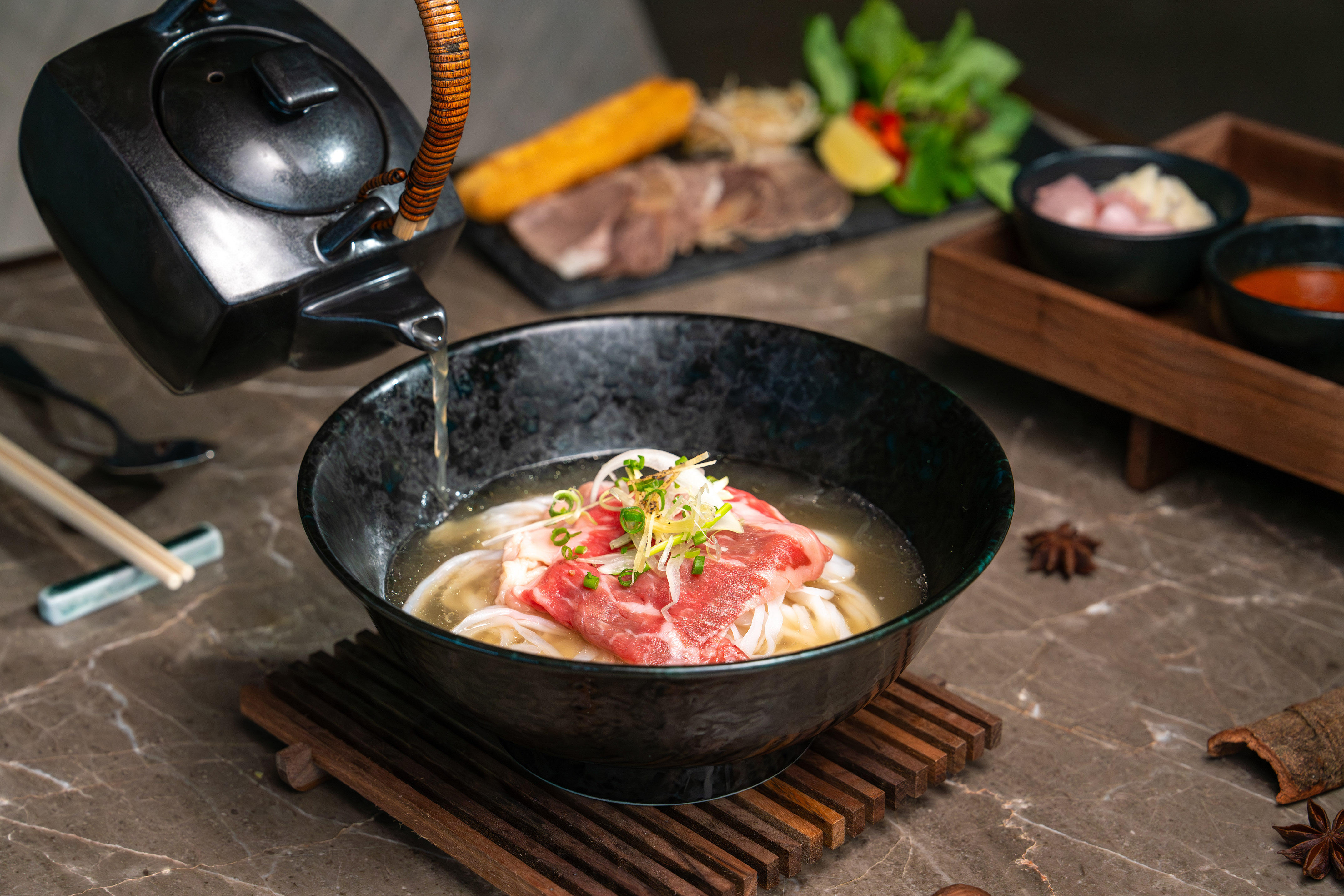 Hot broth poured into beef pho with raw meat, noodles, and fresh herbs.