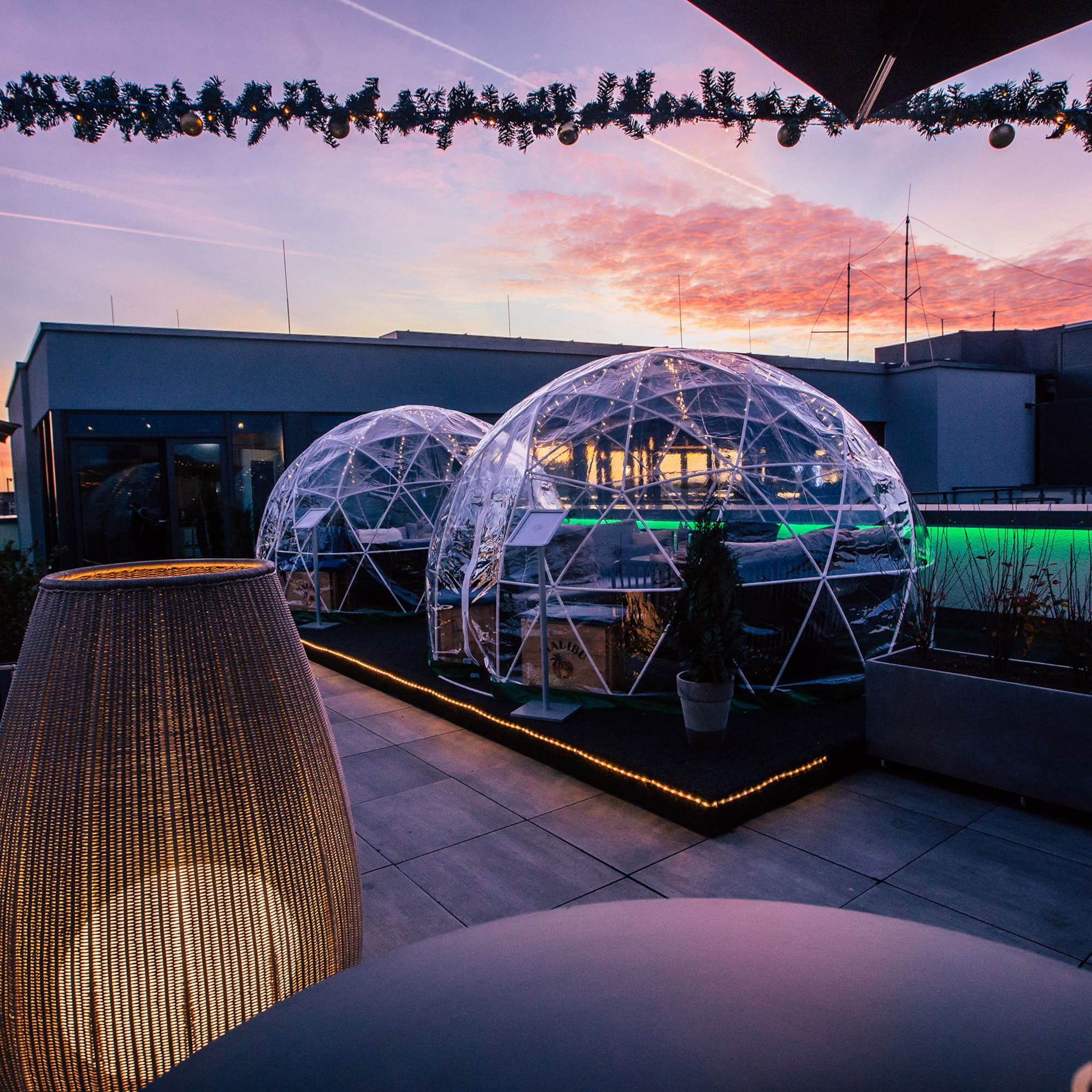 Rooftop terrace with illuminated dining igloos, festive decor, and sunset.