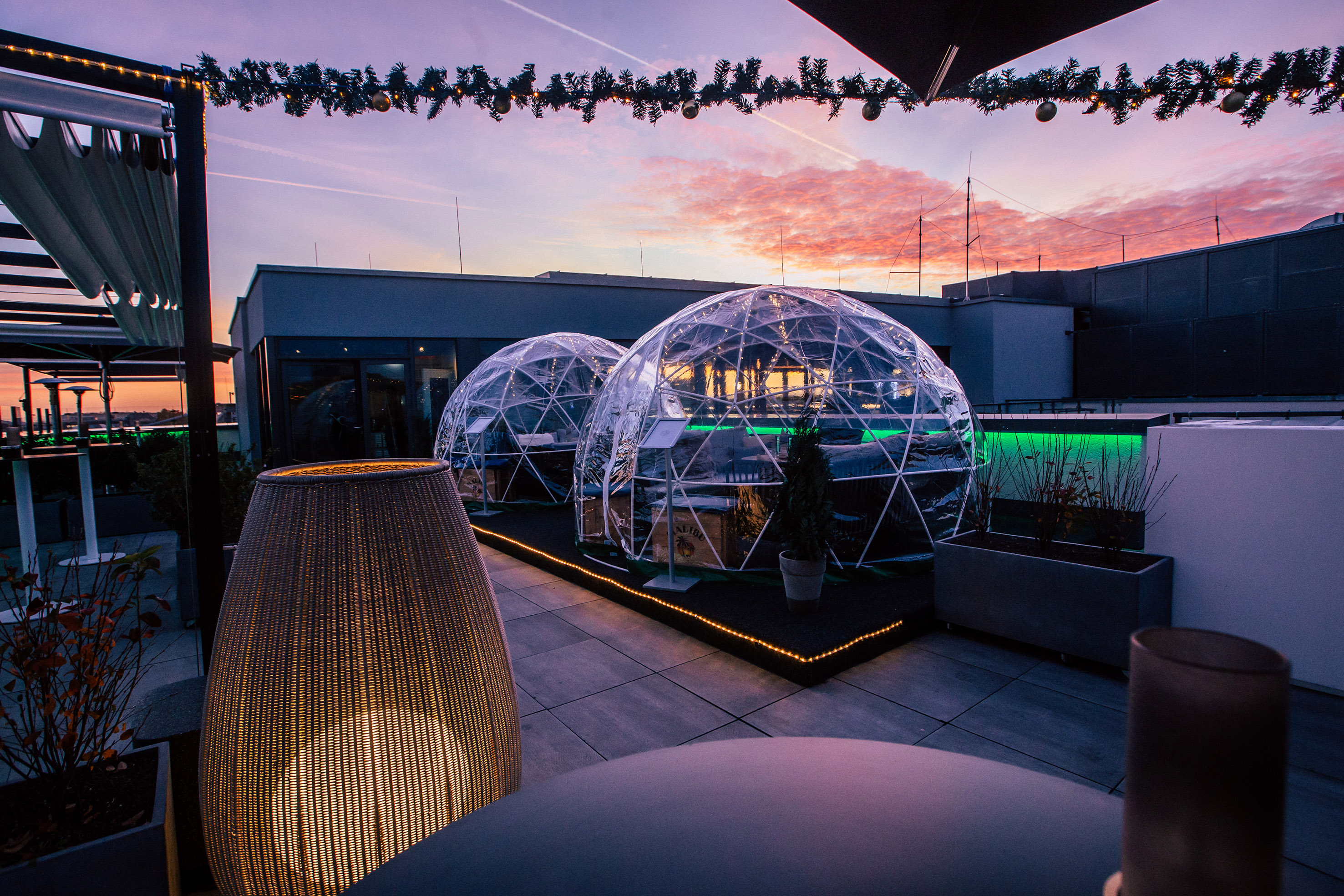 Rooftop terrace with illuminated dining igloos, festive decor, and sunset.
