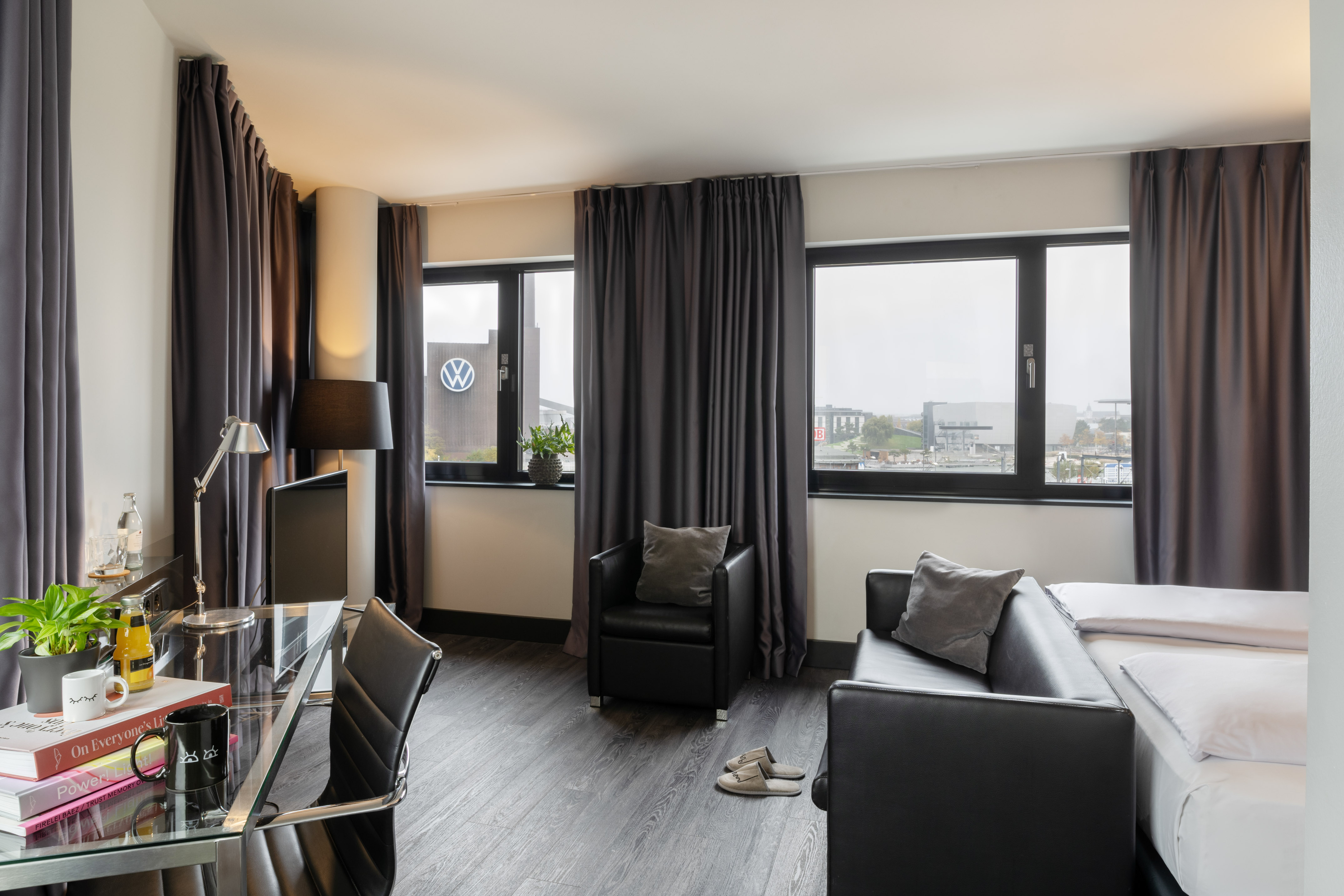 Modern hotel room with desk, seating area, bed, and city view featuring VW logo.