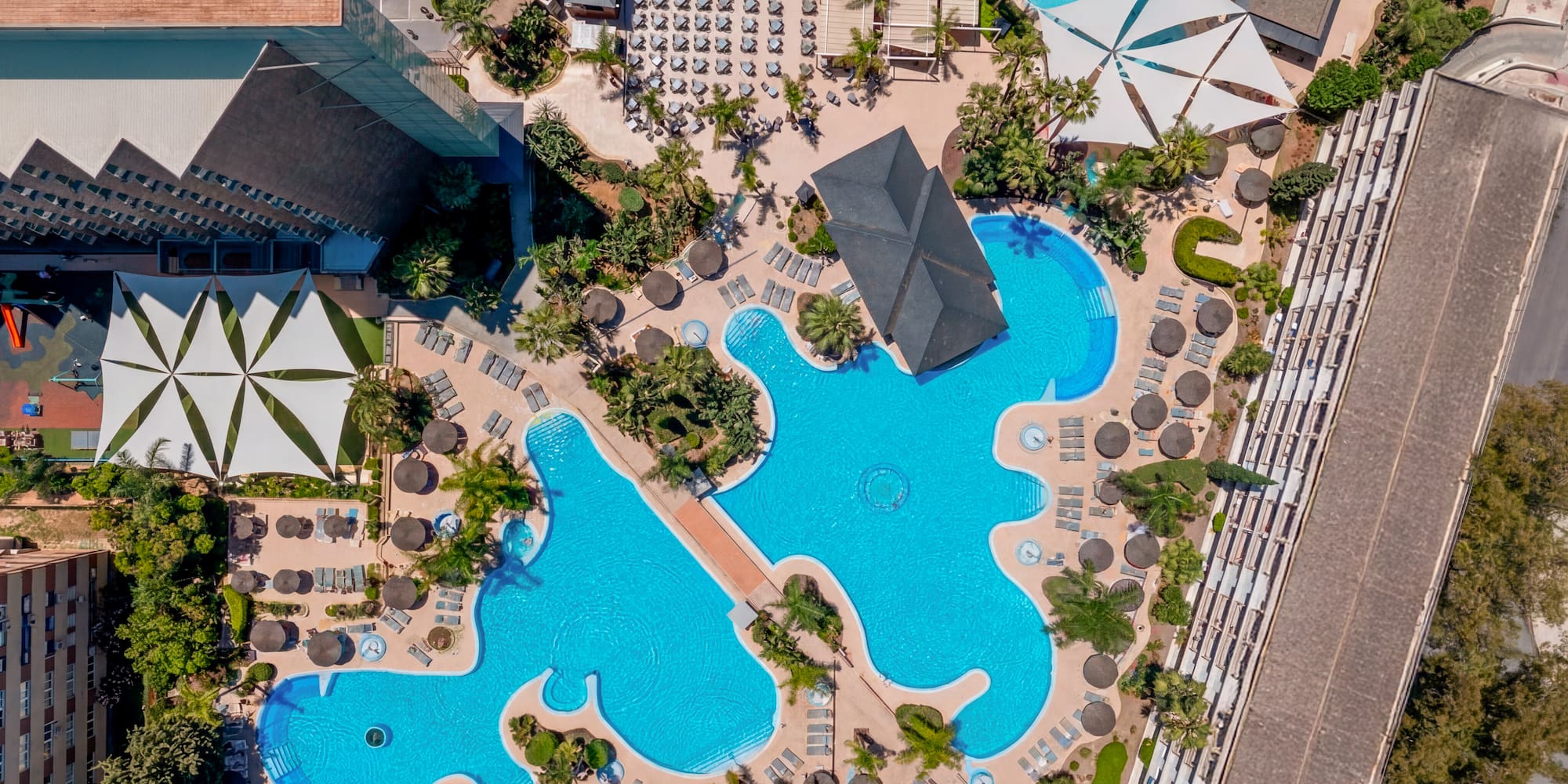Resort aerial: interconnected swimming pools, sun loungers, palm trees, water slides.