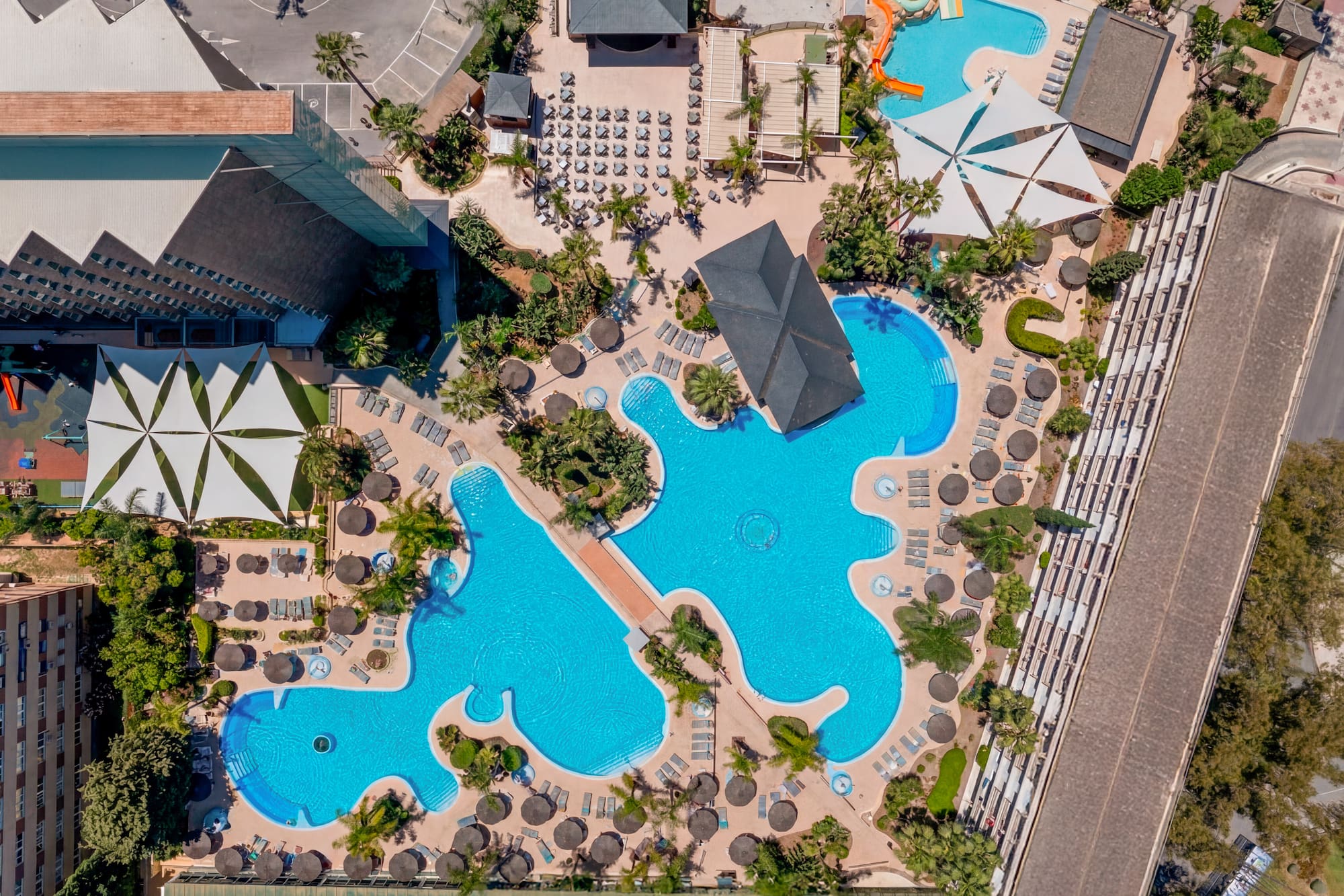 Resort aerial: interconnected swimming pools, sun loungers, palm trees, water slides.
